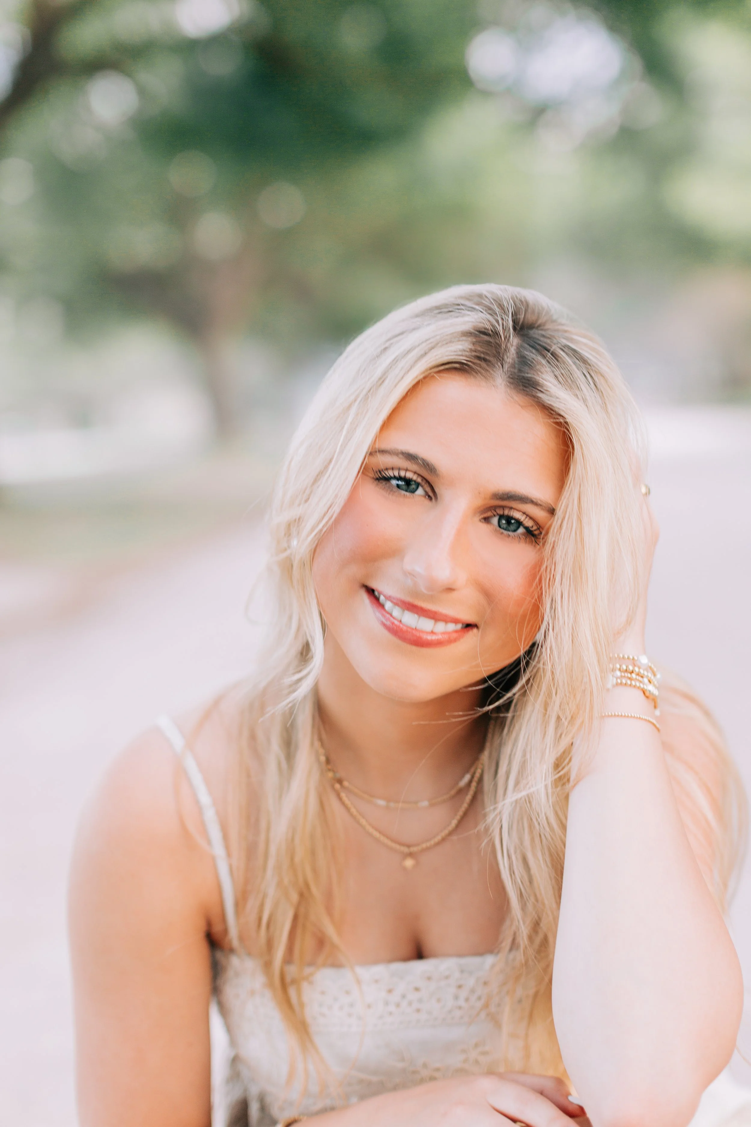 baton rouge senior portraits at burden gardens, blond haired girl sitting on gravel road smiling with trees in background