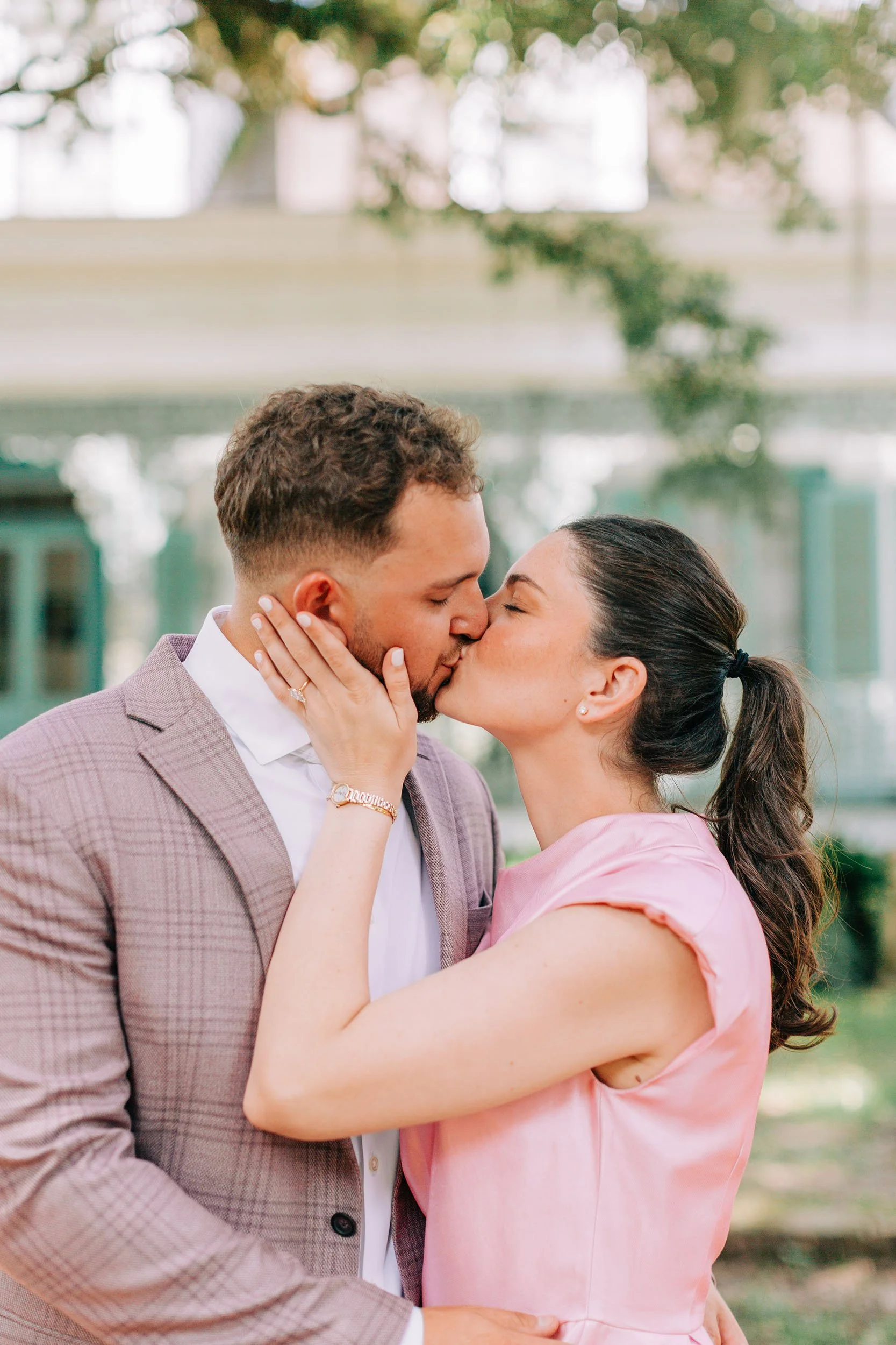 A couple sharing a romantic kiss outdoors, with the woman holding the man's face and the man wearing a light brown suit jacket, white shirt, and a woman in a pink dress with dark hair tied back.