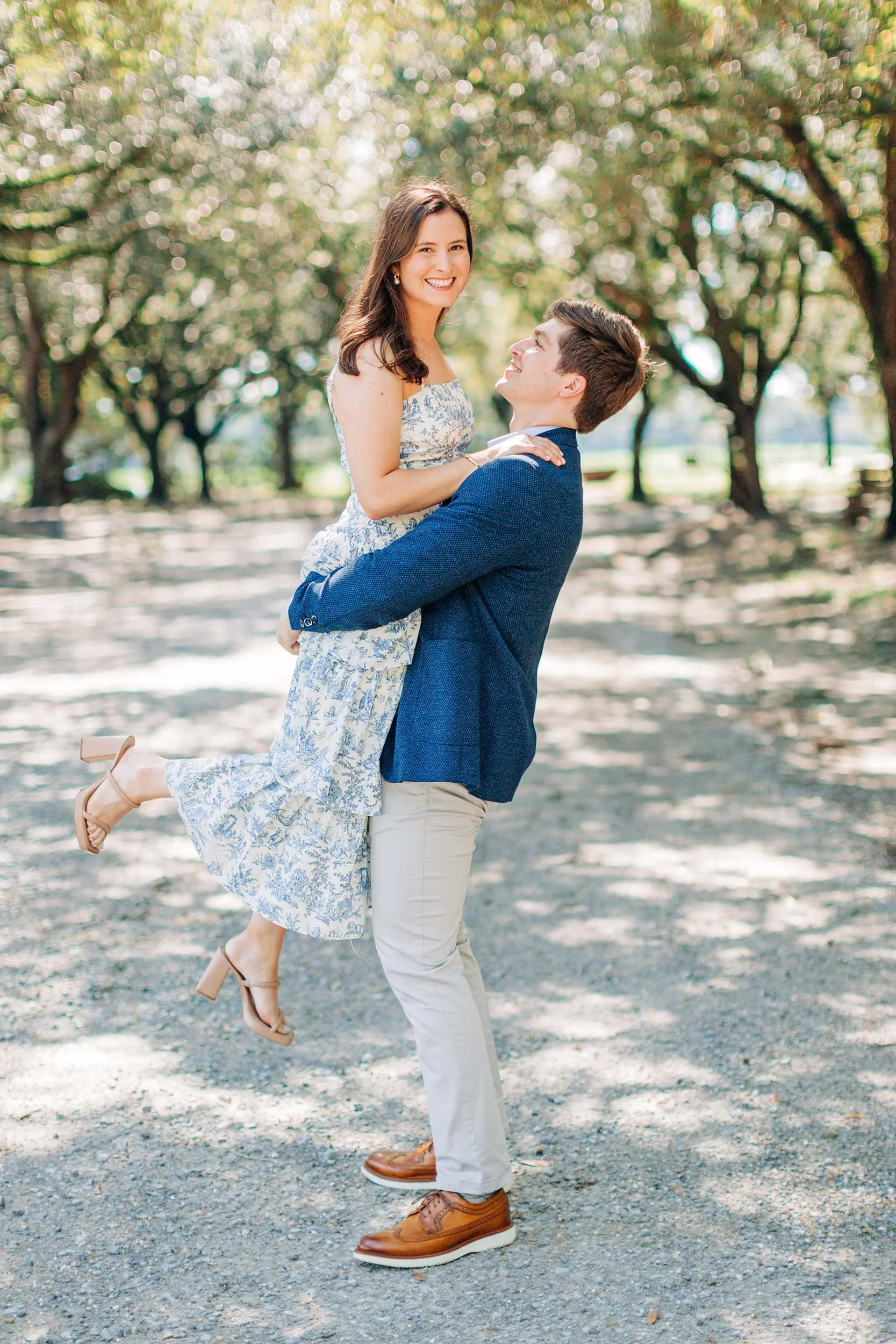 A man is lifting a woman in a park with trees in the background. She is smiling and looking at the camera, while he looks up at her. She is wearing a floral dress and beige high heel shoes, and he is wearing a blue blazer, white pants, and brown shoe