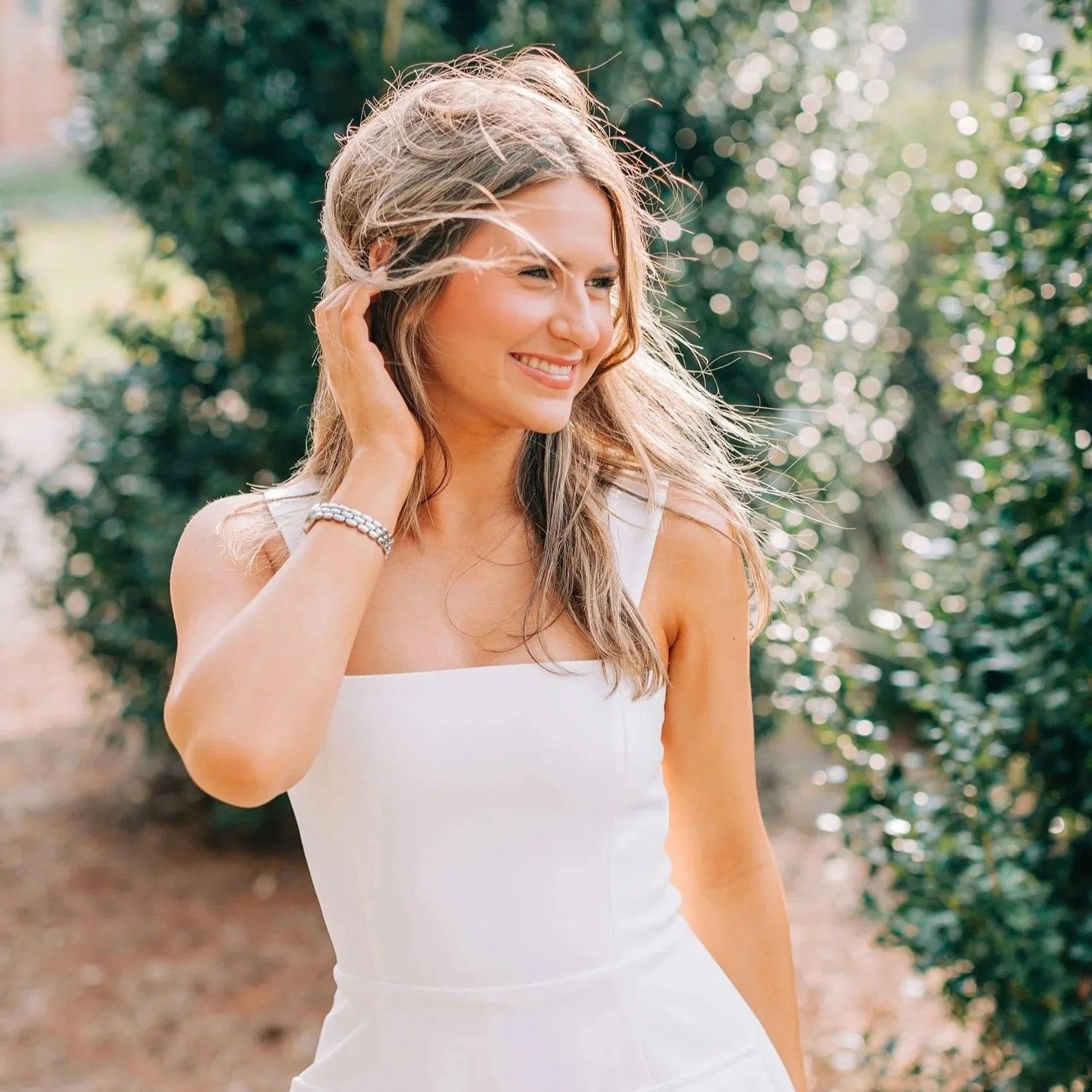 A woman smiling outdoors with sunlight and greenery in the background, wearing a sleeveless white top and a bracelet.