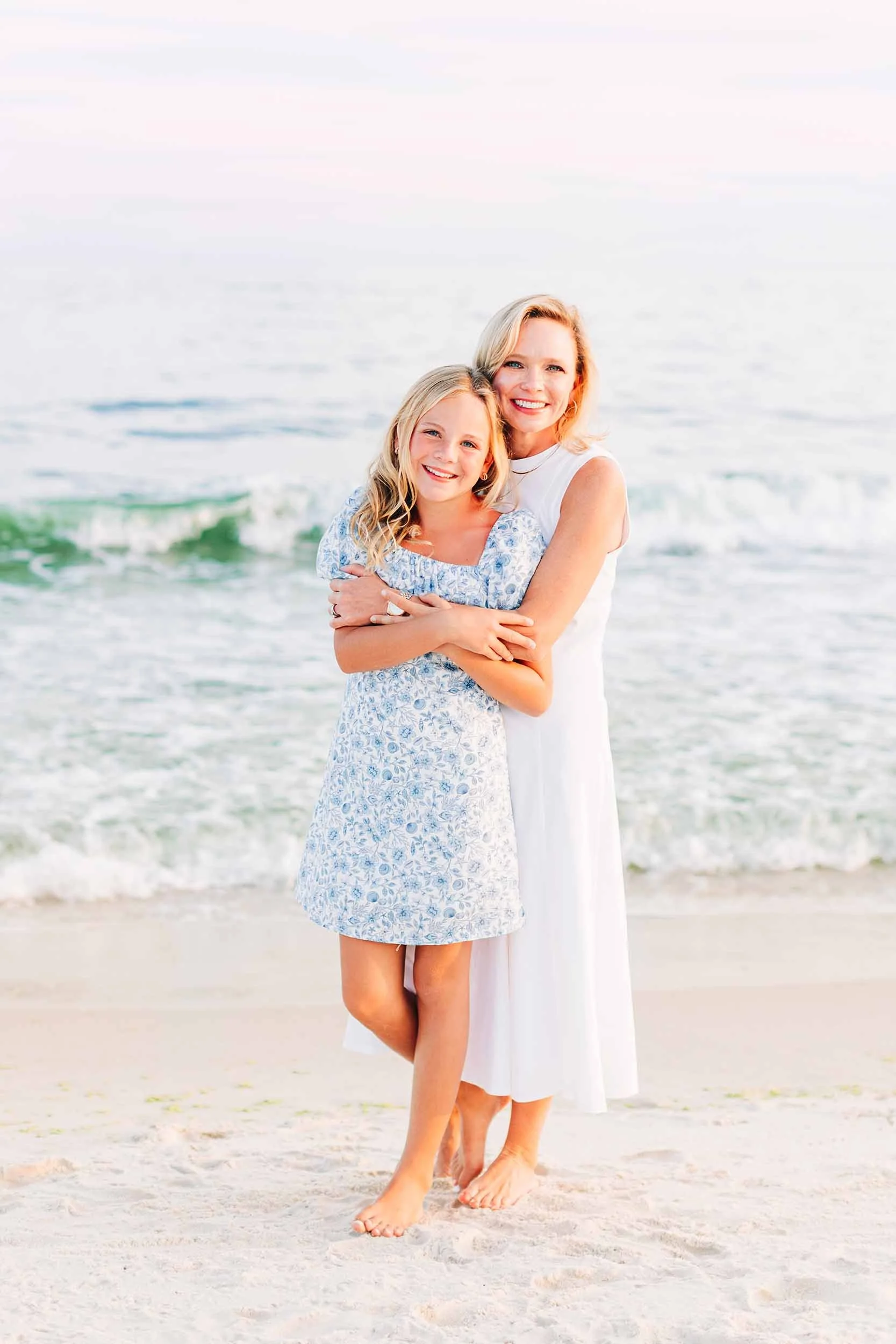 A woman and a young girl smiling on a beach, standing close with arms around each other, with the ocean and waves in the background.