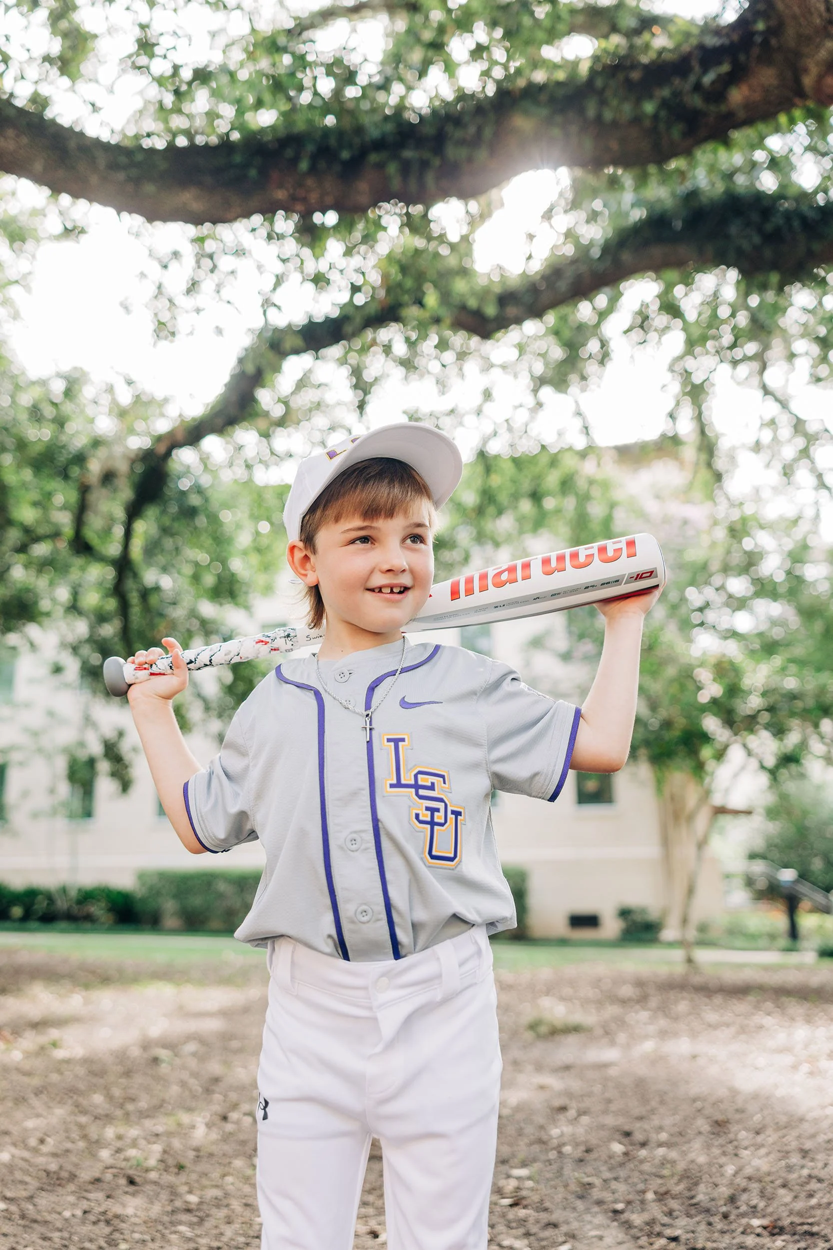 Young boy wearing a baseball uniform holding a baseball bat on his shoulder, standing outdoors under a large tree.
