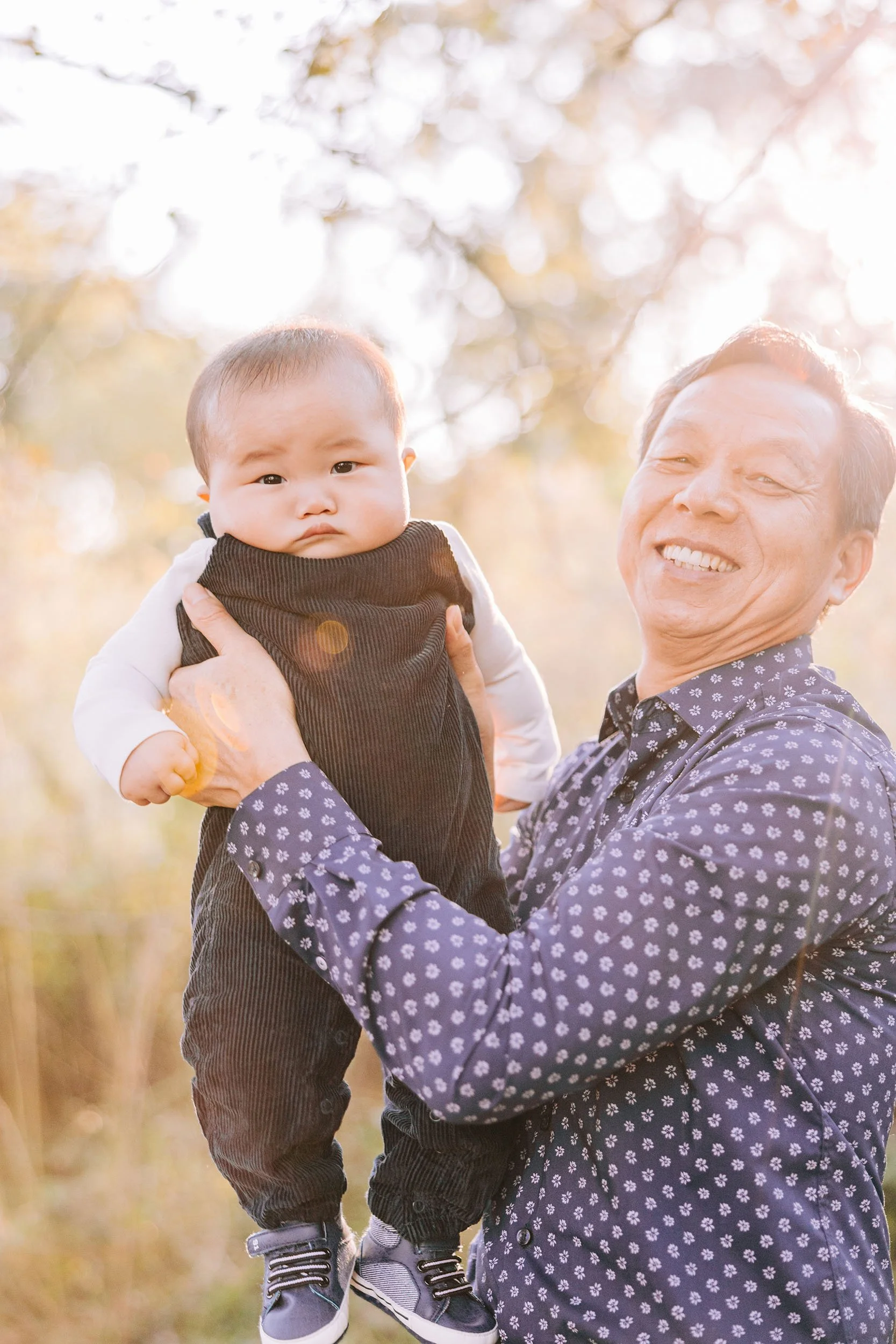 An elderly man with gray hair smiling while holding a young child outdoors during daytime, with sunlight and blurred trees and yellow grass in the background.