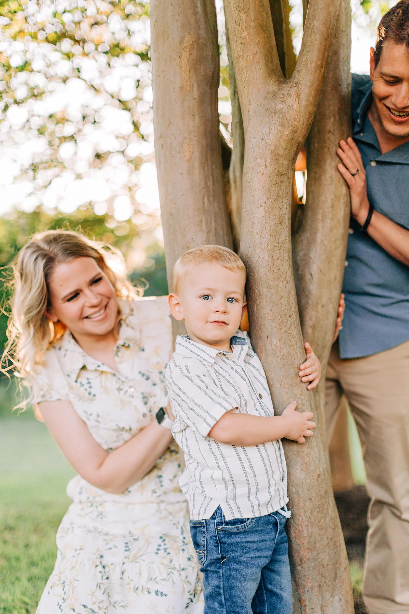 A smiling woman, a young boy with light hair and blue eyes, and a man with a band on his wrist, are playing hide and seek behind a tree in a park.