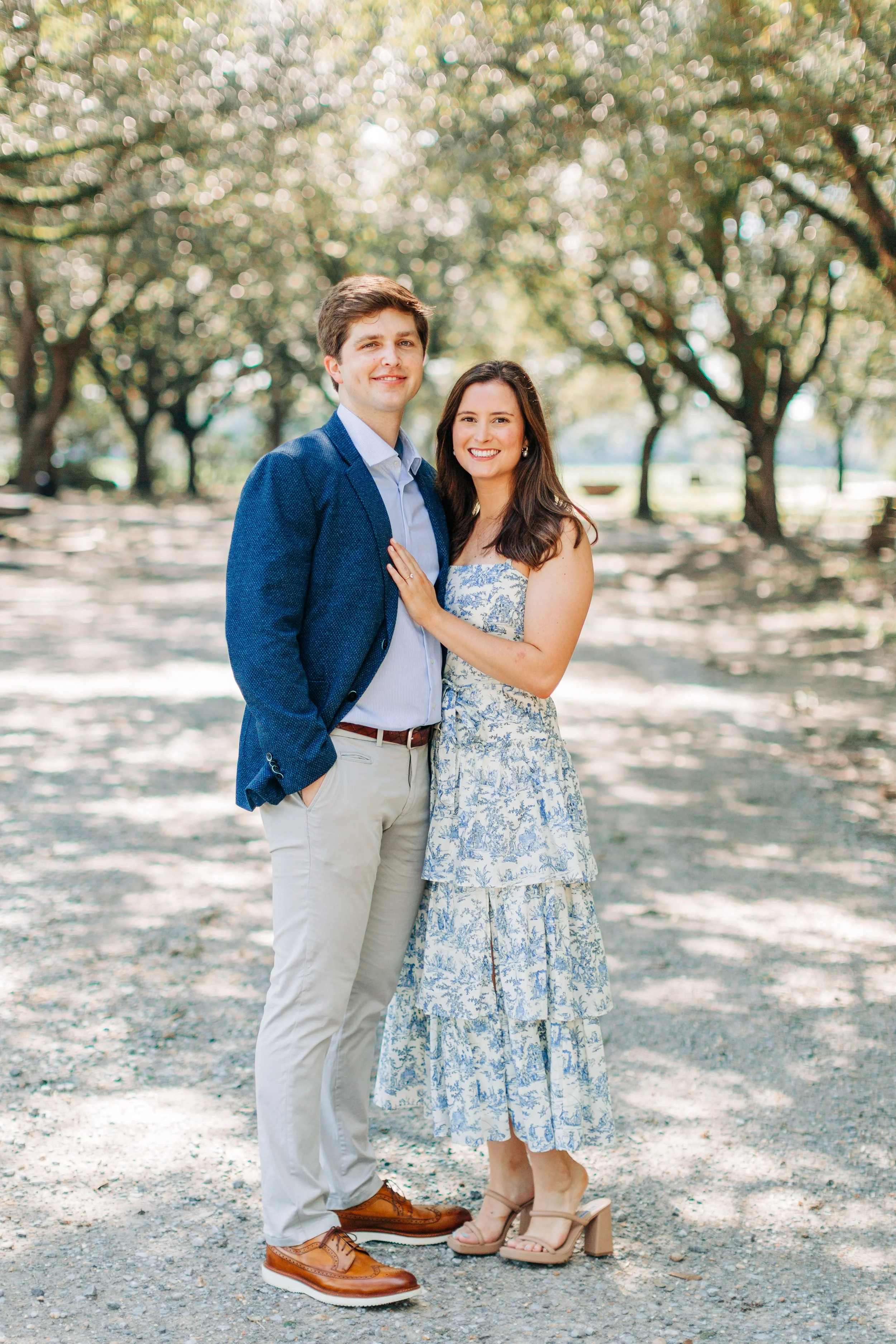A smiling young couple standing outdoors under trees. The man wears a blue blazer, light blue shirt, beige pants, and brown shoes. The woman wears a blue and white patterned dress and beige high-heeled sandals. They are close together, with the woman