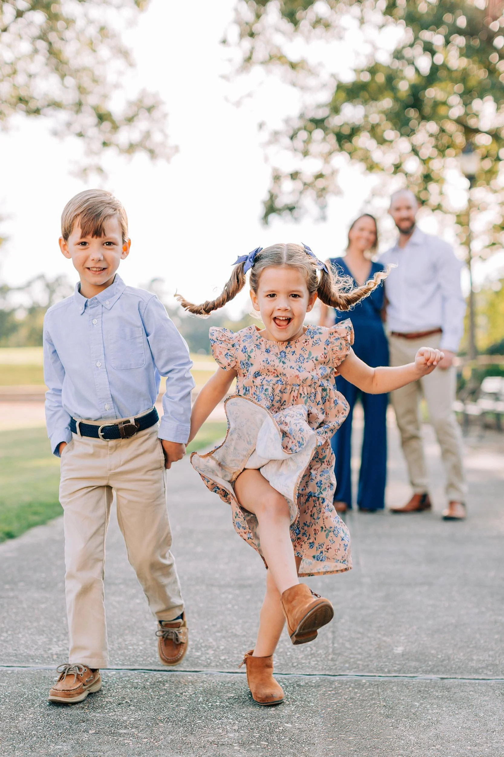 A young girl with pigtails jumping and smiling at the camera, with a boy in a blue shirt and beige pants walking beside her, and a happy family of four in the background outdoors on a sunny day.
