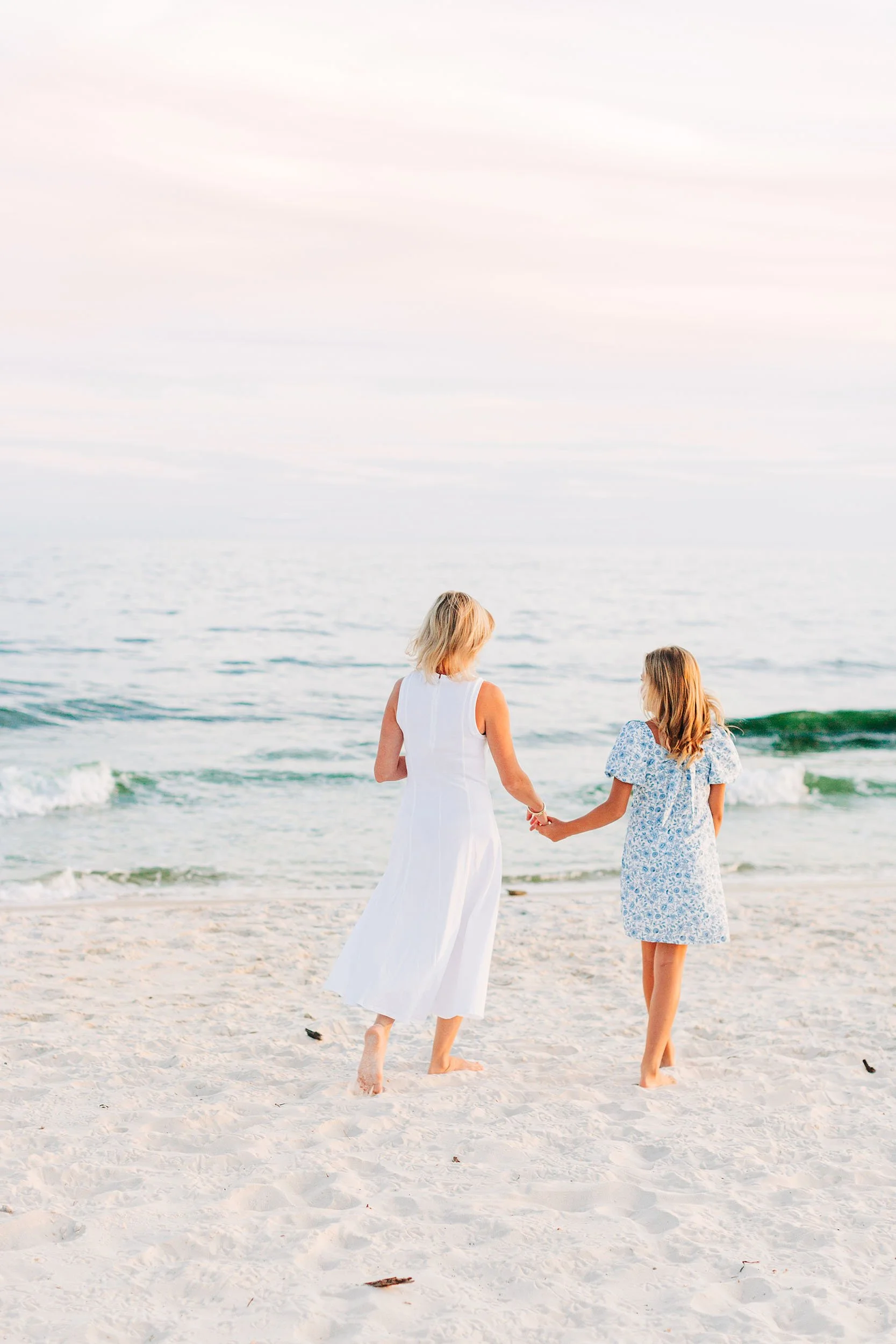 A woman and a young girl hold hands and walk along the beach at sunset, with the ocean waves in the background.