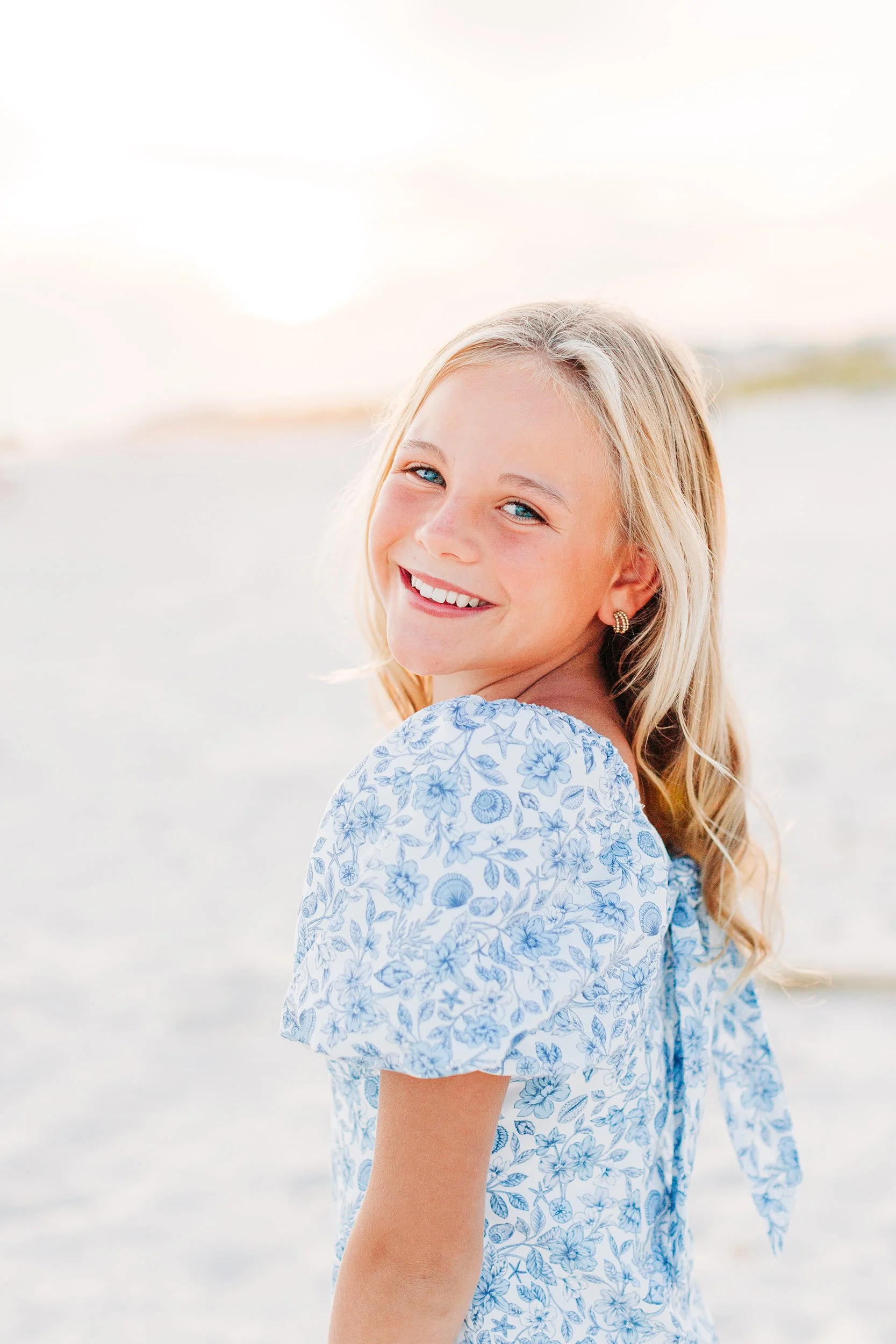 Close-up of a smiling young girl with blonde hair, blue eyes, and earrings, wearing a white dress with blue floral patterns, standing outdoors near a beach at sunset or sunrise.