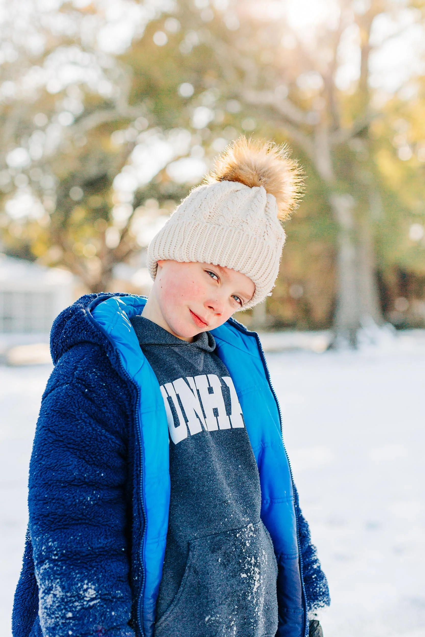 A young boy standing outside in winter clothing, including a cream knit hat with a fur pom-pom, a dark gray hoodie, and a blue jacket, with snow on his hoodie. He is smiling slightly with a snowy background and leafless trees.