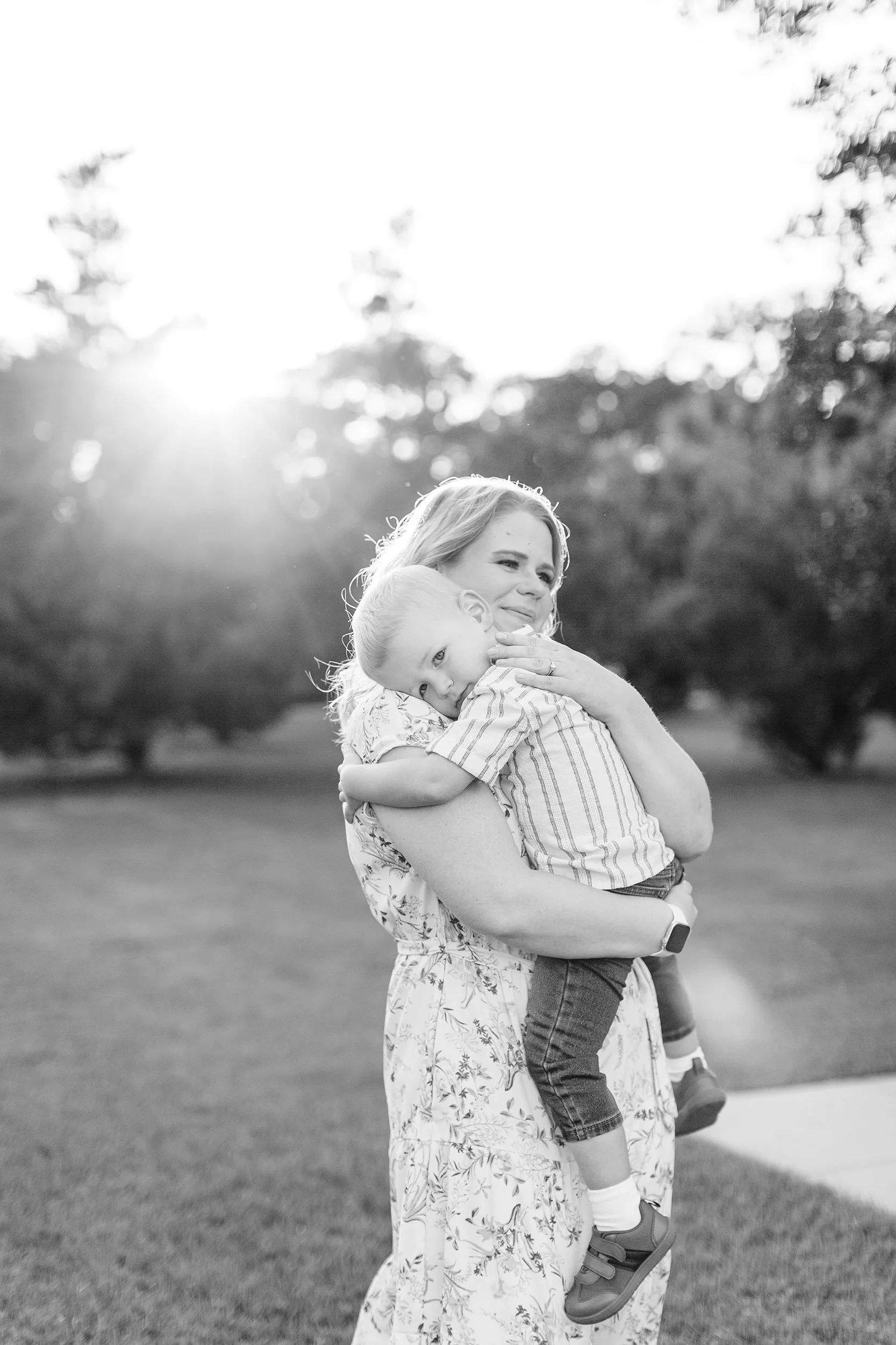 A woman with light hair holding a young boy, with trees and sunlight in the background, in black and white.