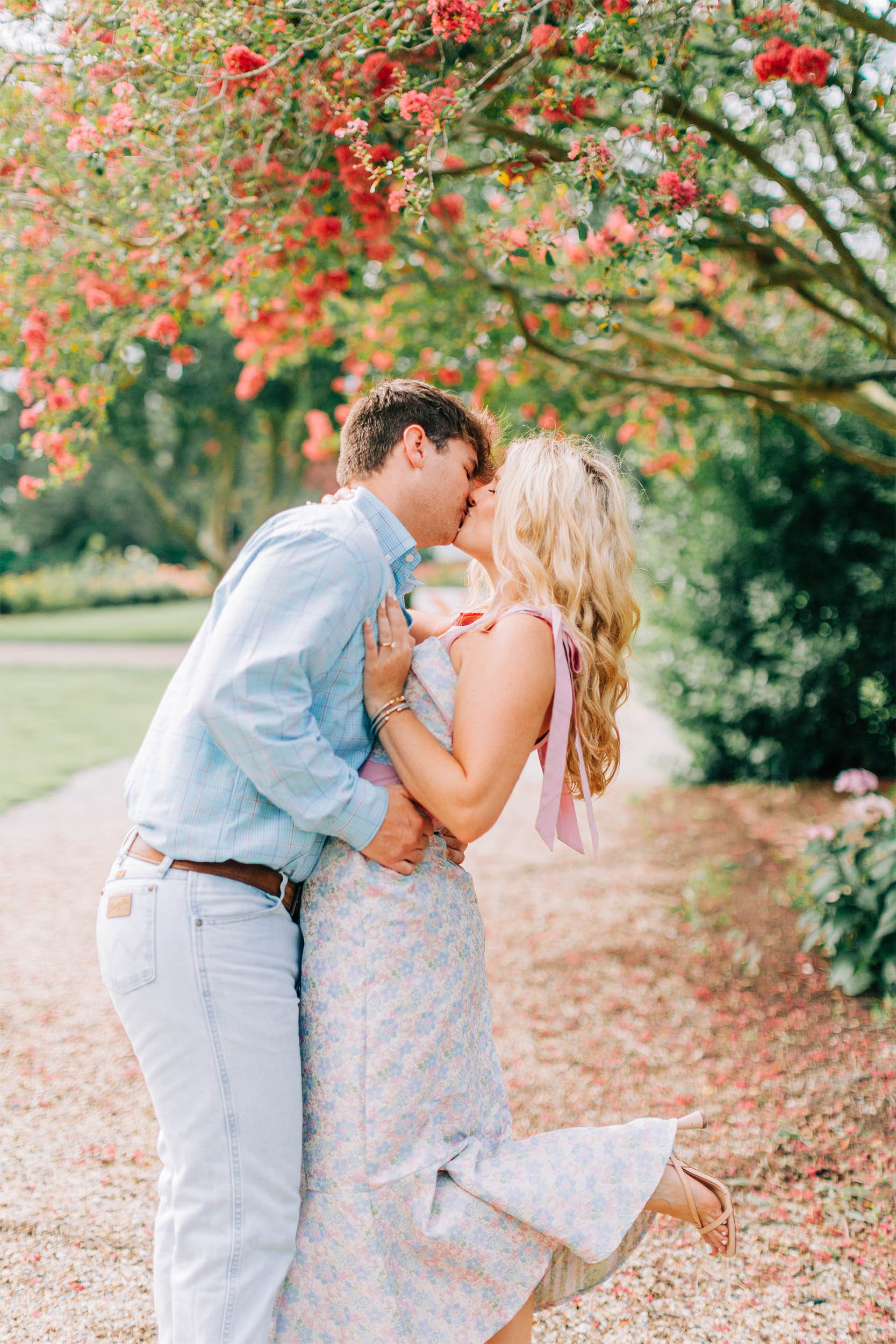 A young couple is kissing outdoors under a flowering tree with pink and red blossoms. The woman, with curly blonde hair, is wearing a floral dress and sandals, while the man is in a light blue checkered shirt and white pants. She is standing with her