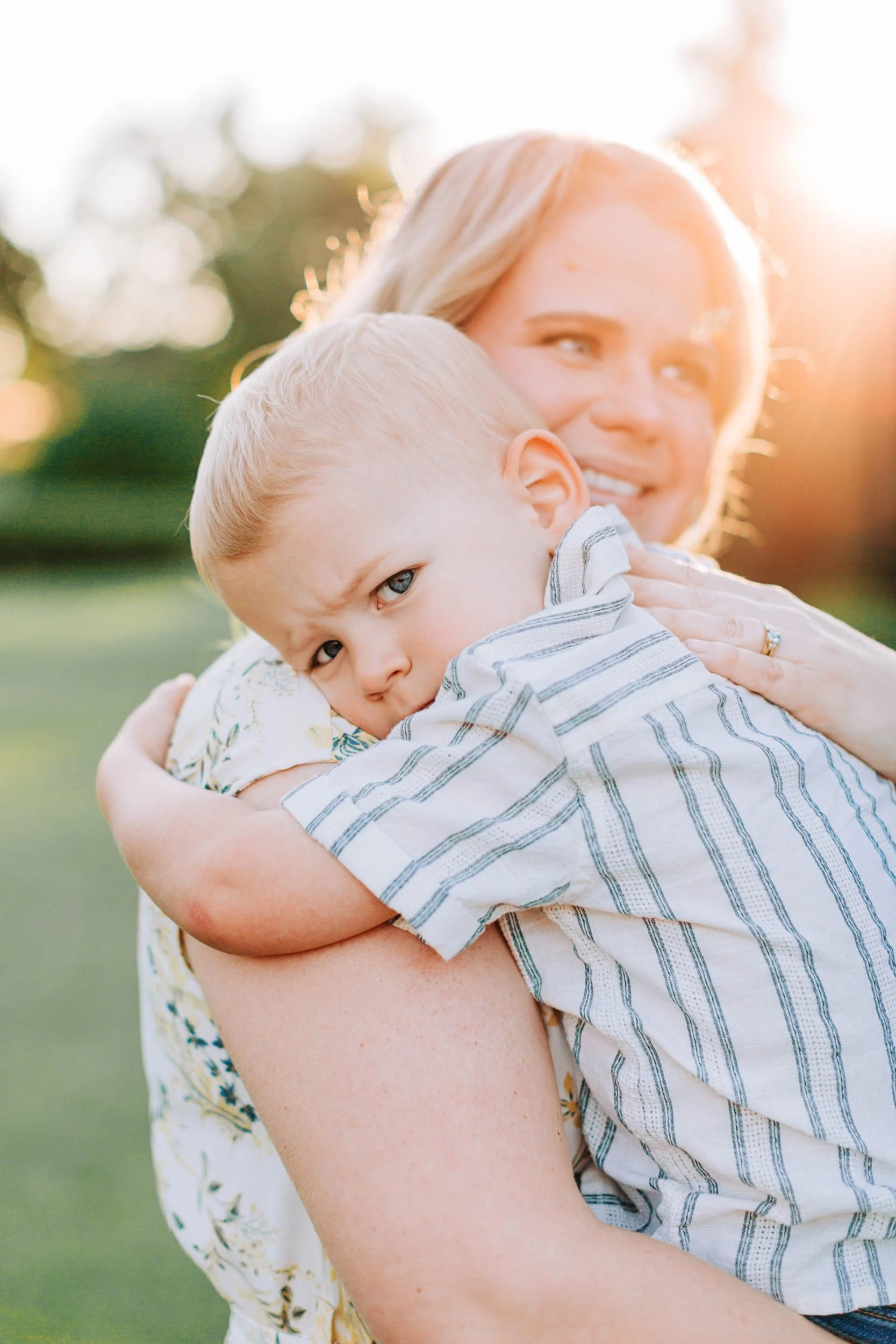 A woman is hugging a young boy outdoors during sunset. Sunlight creates a warm glow around them, and they are standing on a grassy area with blurred trees in the background.