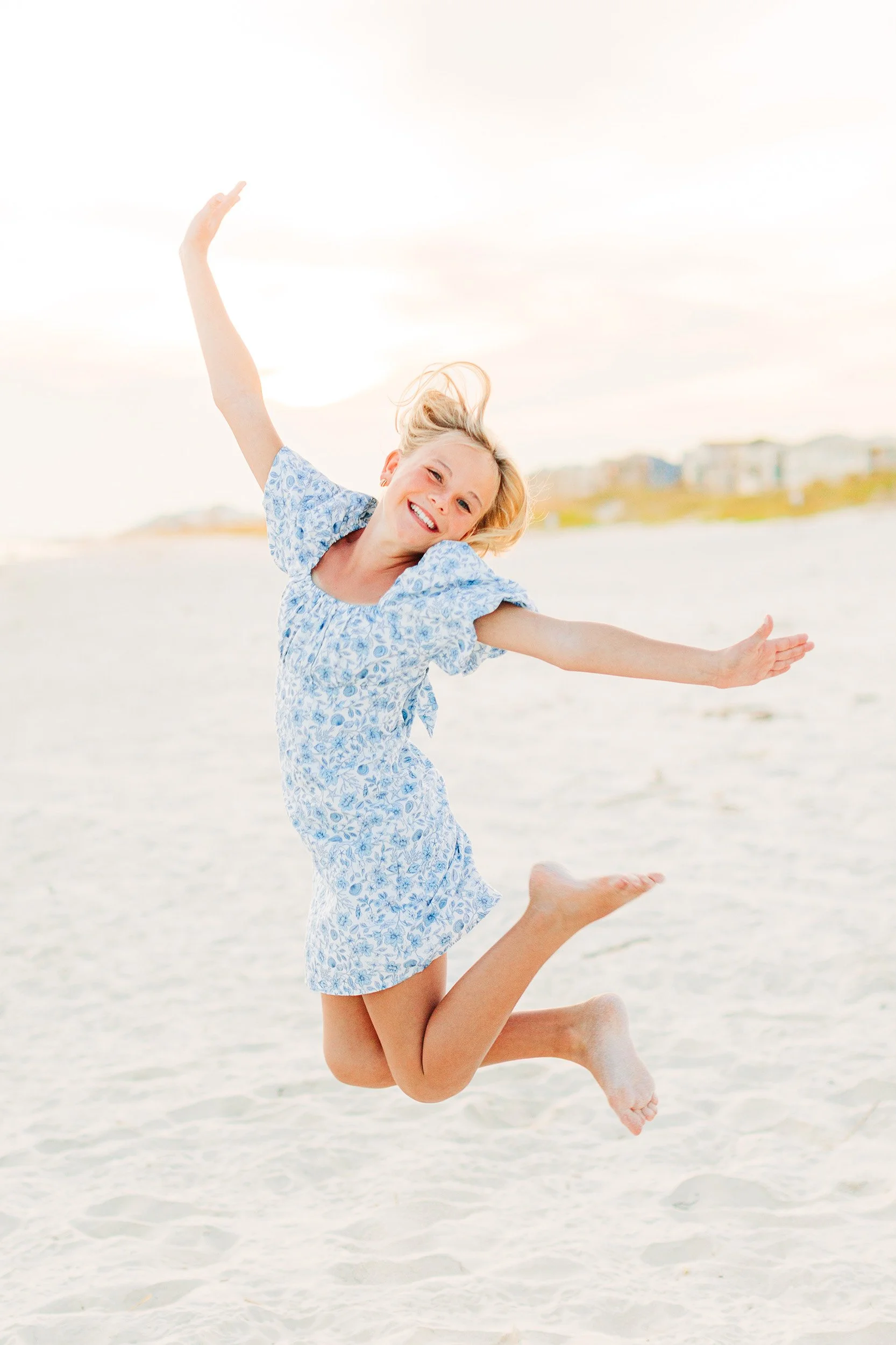 A young girl jumping on the beach with arms outstretched and a big smile, wearing a blue and white floral dress, with houses in the background under a light sky.