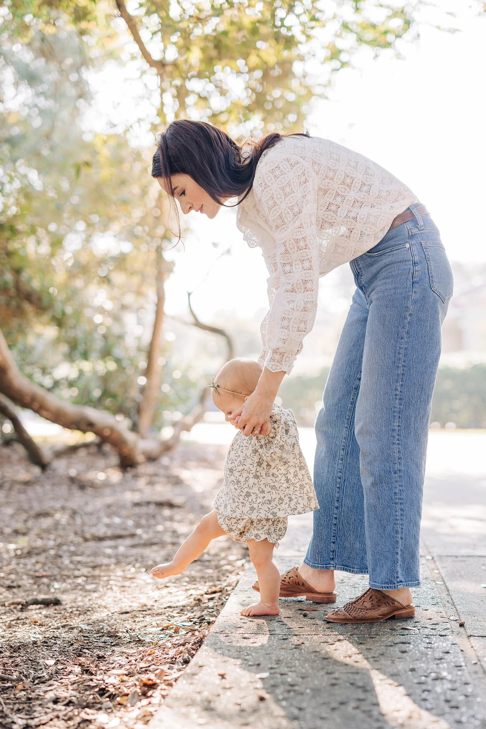 A woman with dark hair wearing a white lace blouse and jeans helping a young girl in a beige floral dress learn to walk, outdoors on a sunny day with trees in the background.