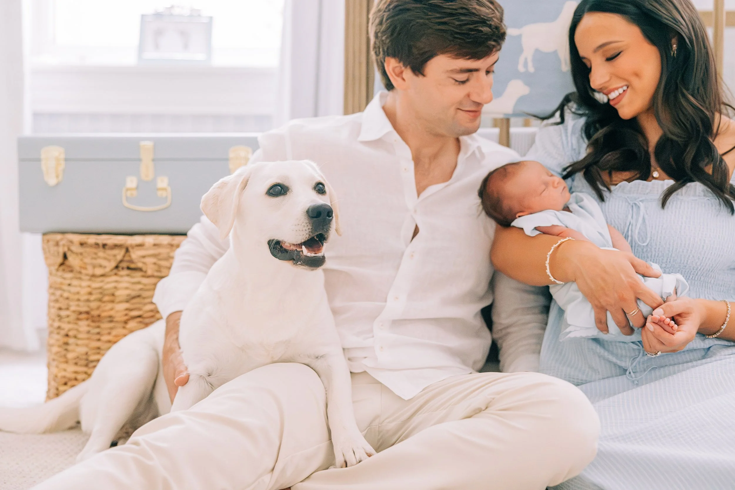 A man and woman sitting happily on a beige sofa with a Labrador retriever puppy and a newborn baby. The man is wearing a white shirt, the woman has long dark hair, and they are near a window with a blue wall decor featuring white animal silhouettes.