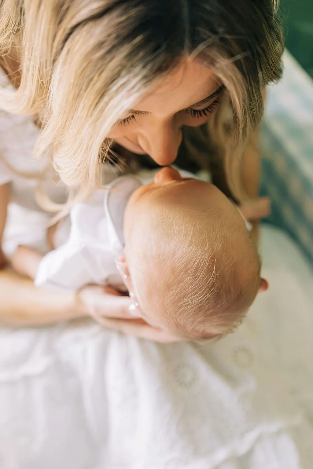 A woman with blonde hair gently kisses the forehead of her baby, who has light-colored hair, while lying on a bed or changing table.