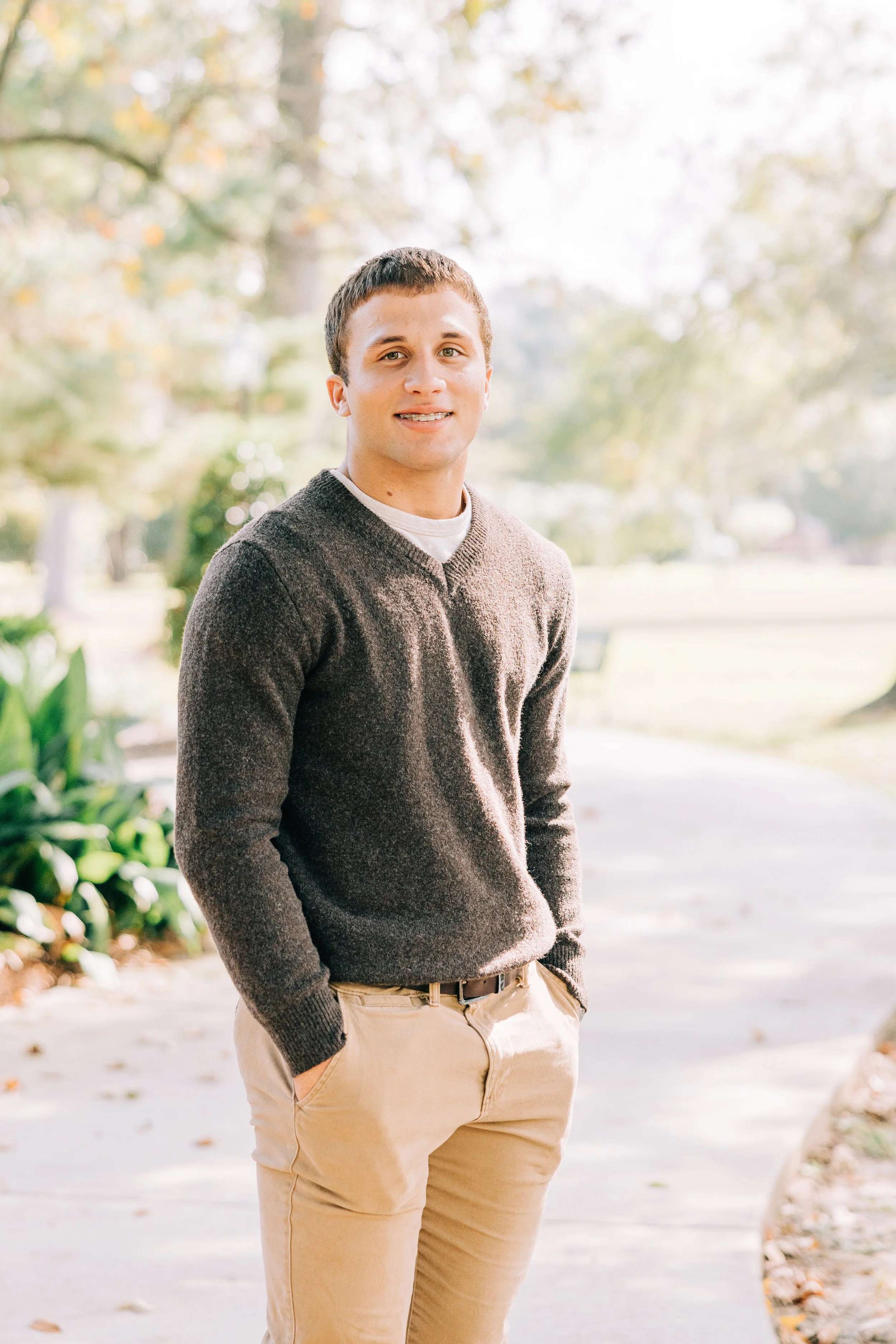 A young man with light skin and short brown hair standing outdoors in a park or garden, wearing a brown sweater and beige pants, with trees and sunlight in the background.
