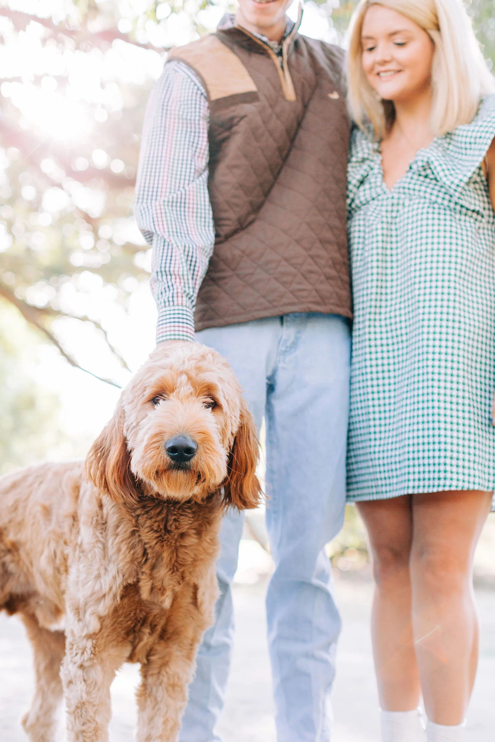 A golden retriever dog standing outdoors with two people, a man and a woman, behind it. The dog has a fluffy coat and dark eyes, looking directly at the camera. The man is wearing a brown vest over a checkered shirt, and the woman is wearing a blue a