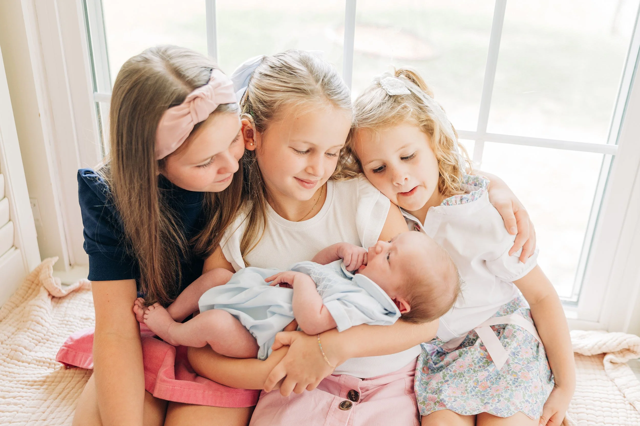 Four young girls hugging and looking at a newborn baby on a bed near a window.