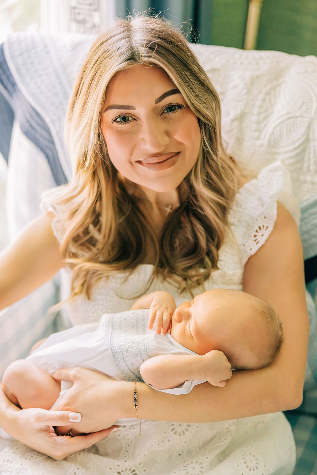 A woman with long blonde hair holding a sleeping baby inside a well-lit room.