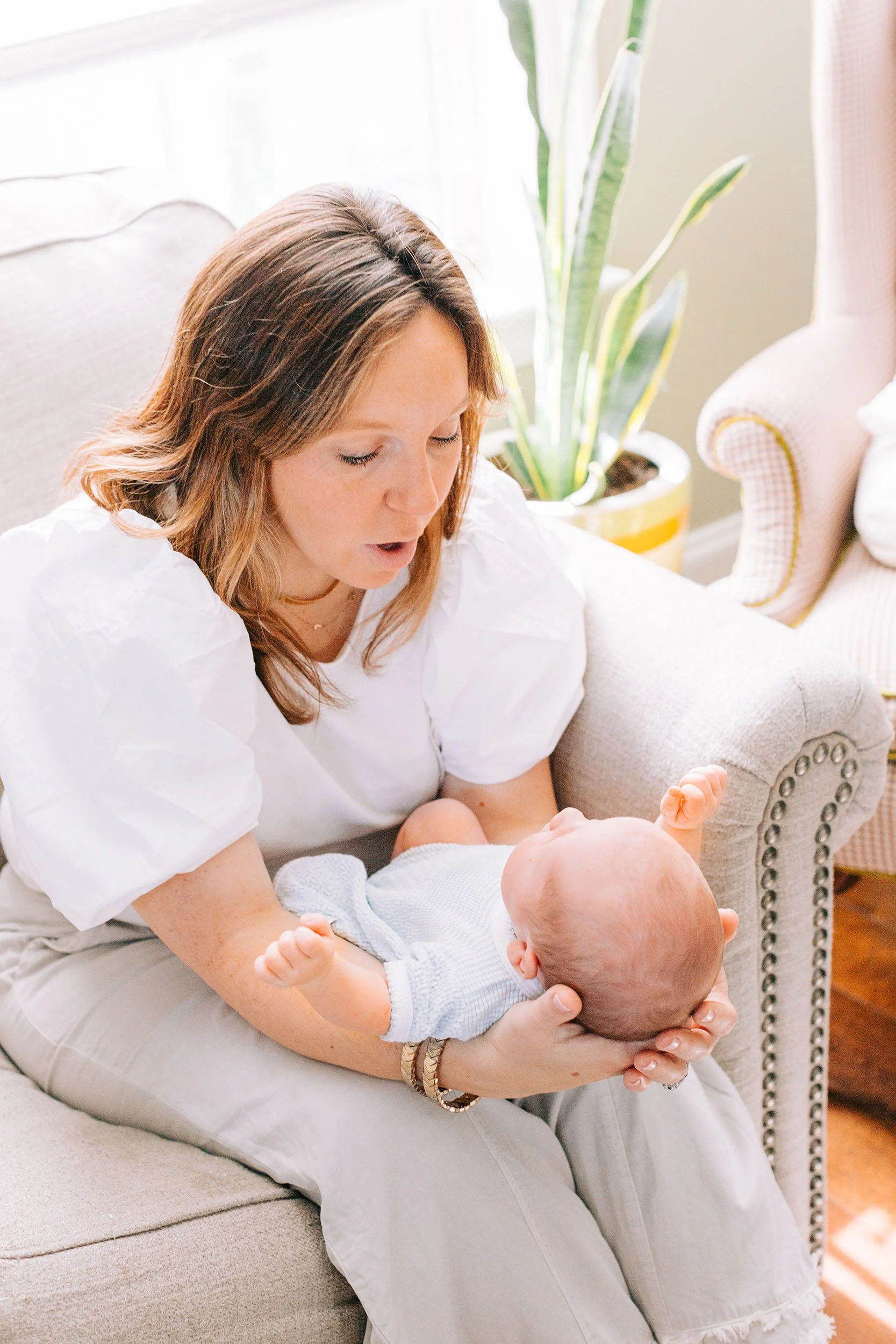 A woman with brown hair and wearing a white shirt holds a baby, who is lying on her lap, in a bright living room with plants and furniture.