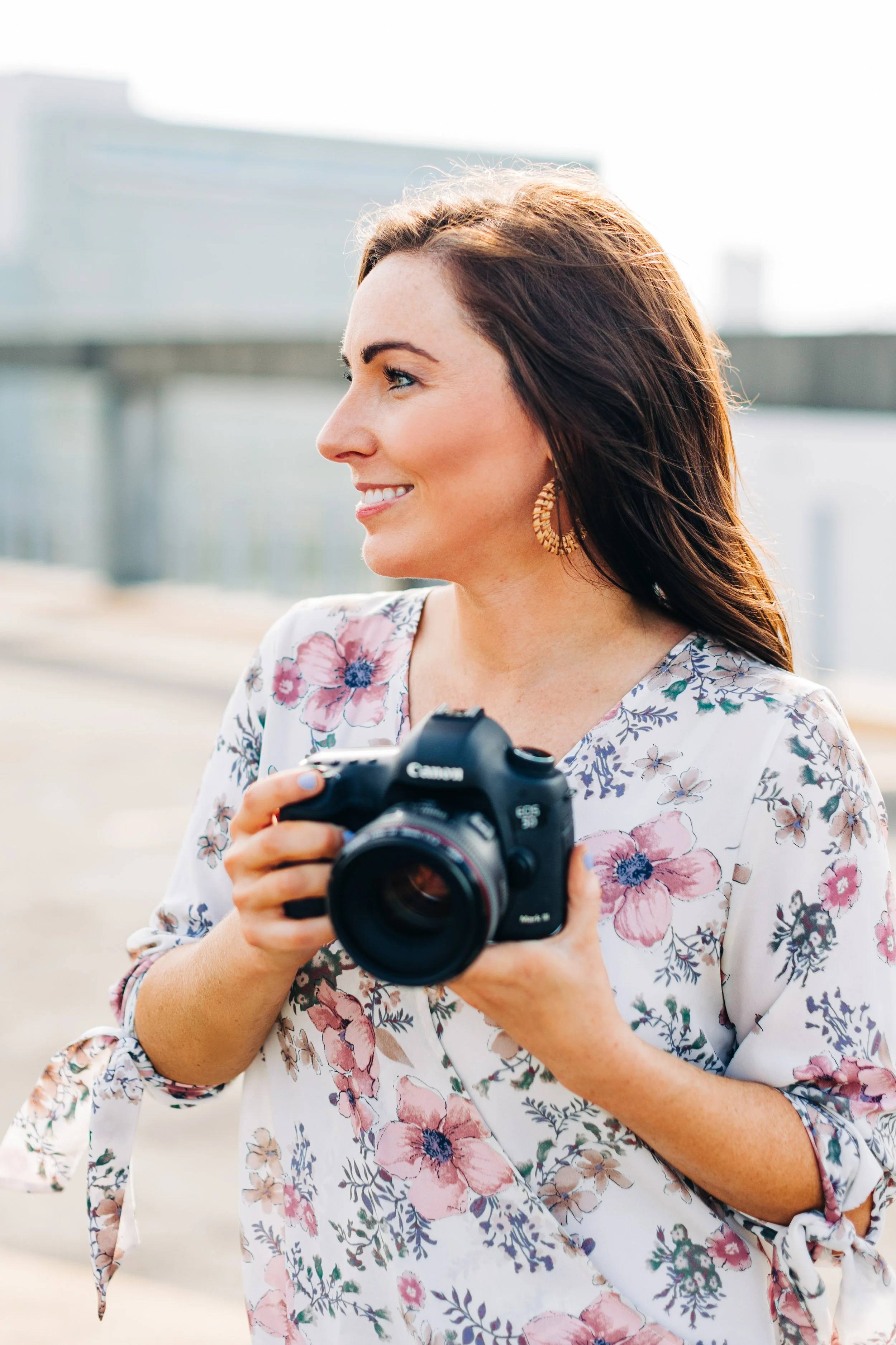 A woman with long brown hair, smiling, wearing a floral blouse and hoop earrings, holding a Canon camera outdoors near a body of water with a bridge in the background.