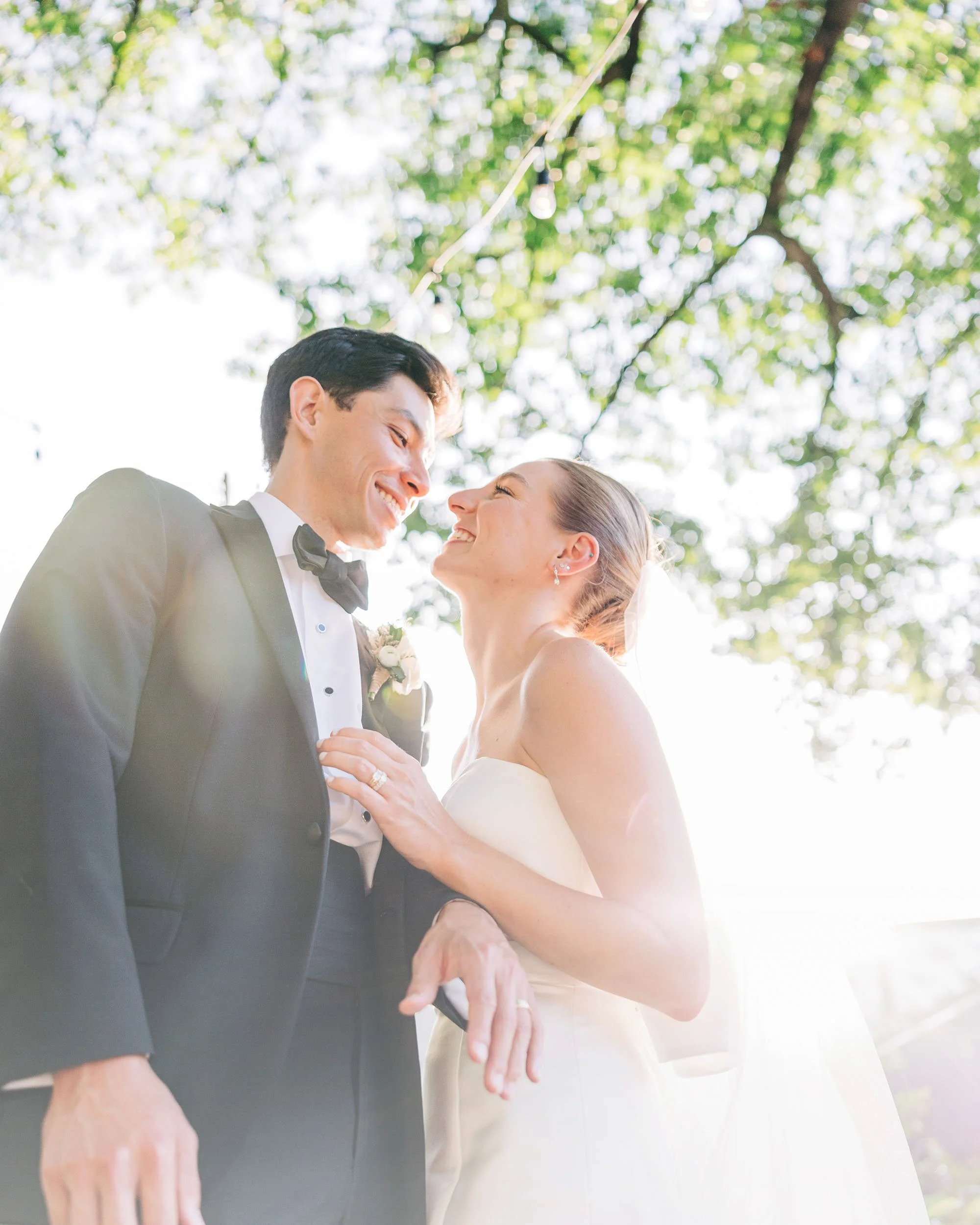 Close-up of a happy couple in wedding attire, holding hands outdoors with green trees and sunlight in the background.