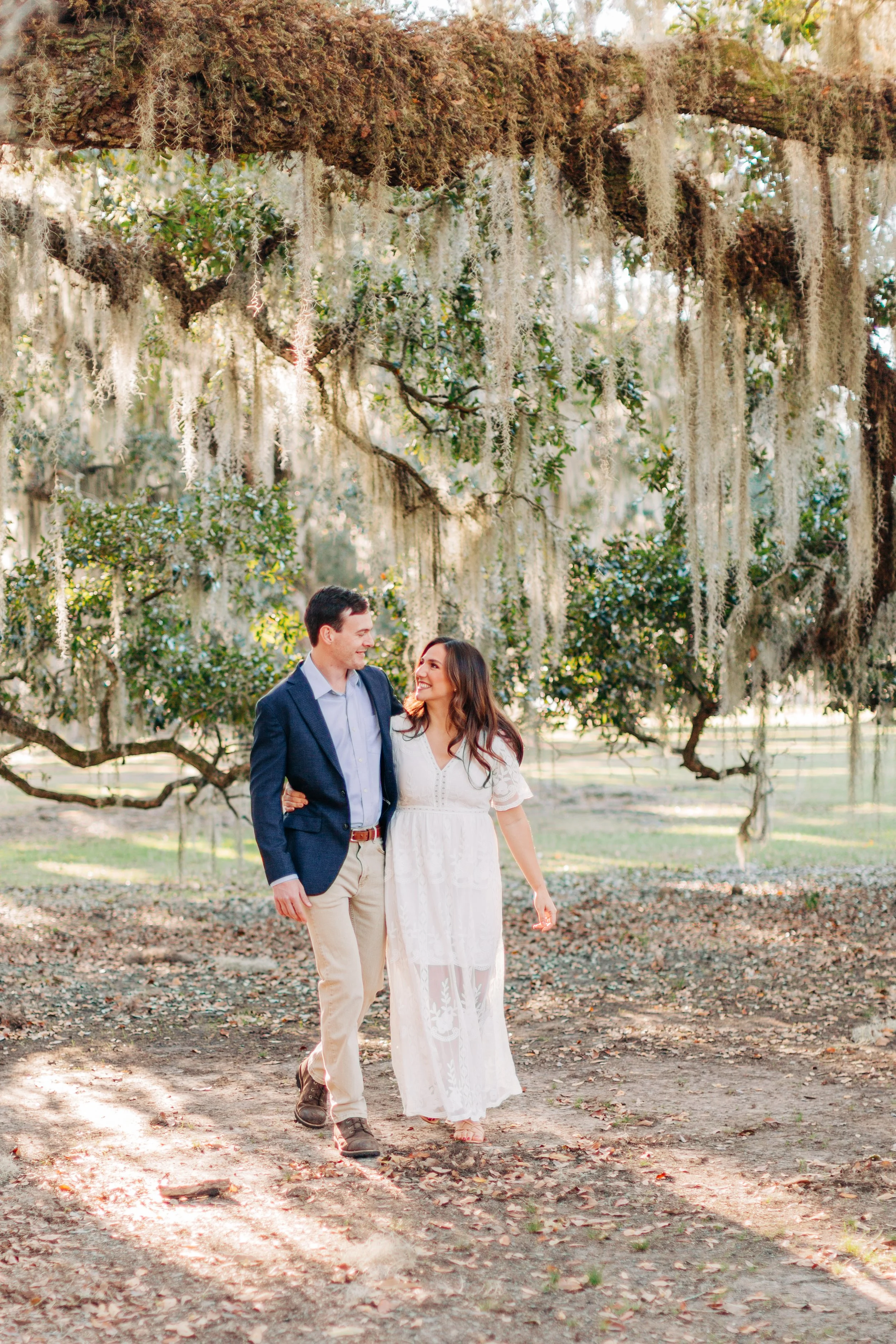 A couple walking in a park under a large tree with Spanish moss, sunlight filtering through the branches, leaf-covered ground, the man is wearing a blazer and khakis, the woman is in a white dress, smiling and looking at each other.
