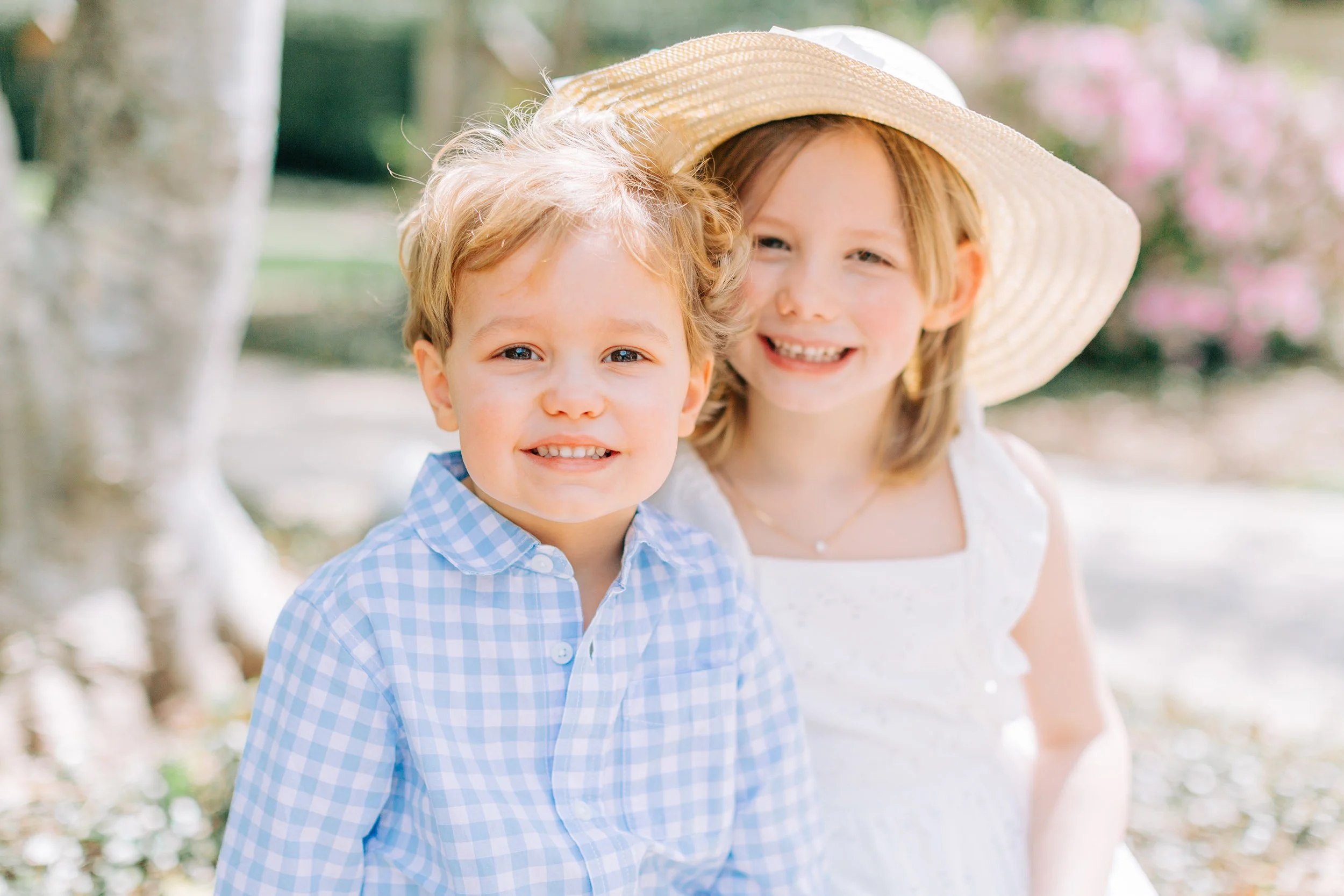 A young boy and girl smiling outdoors, with the girl wearing a large straw hat and the boy in a blue checkered shirt.