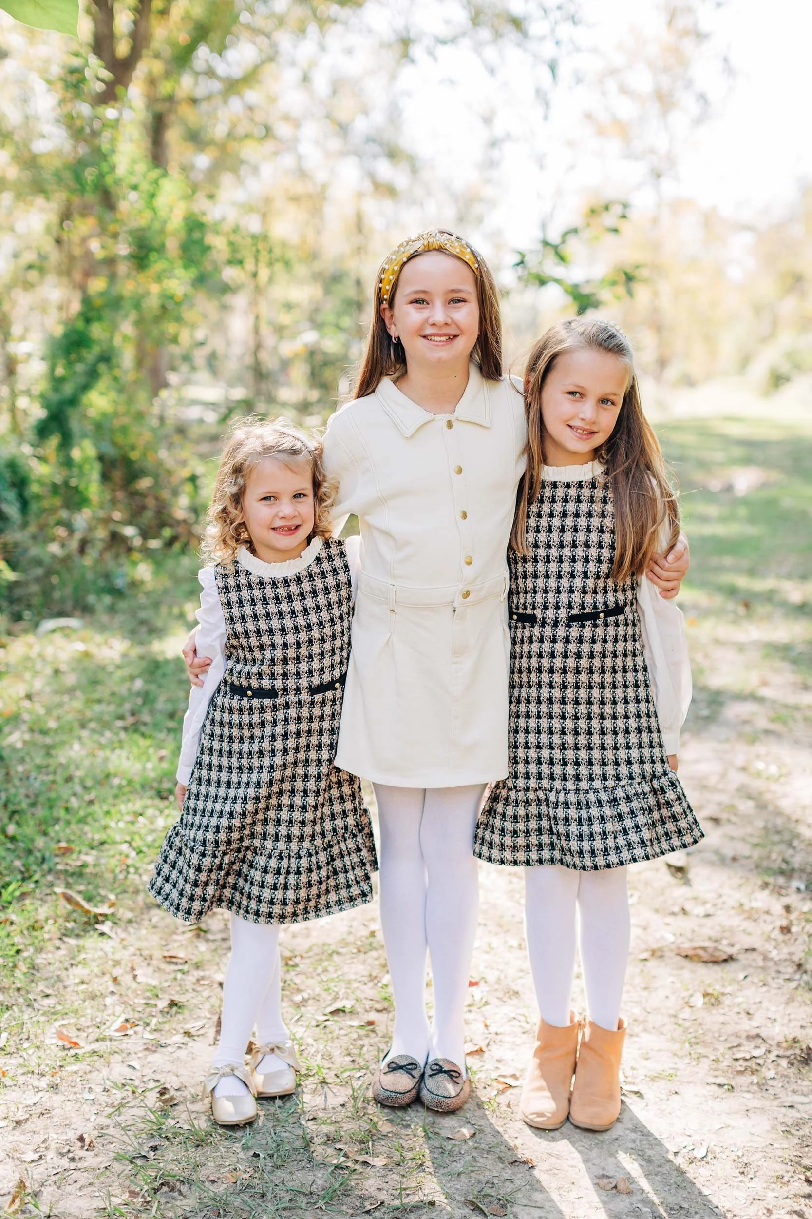 Three young girls standing together outdoors on a sunny day, wearing matching black-and-white patterned dresses with white tights and shoes, smiling at the camera.