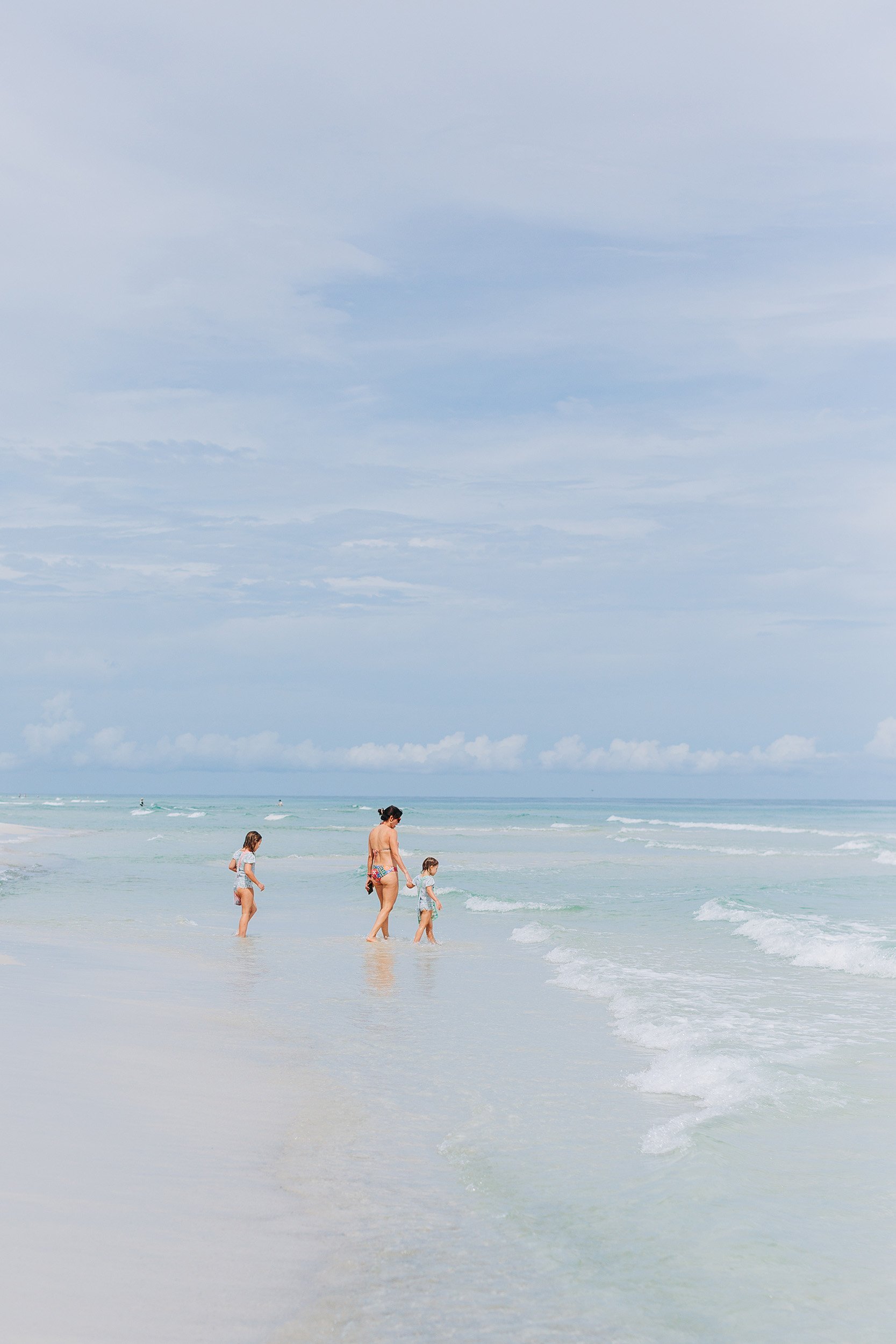 A woman and two children walking in shallow ocean water at the beach on a cloudy day.