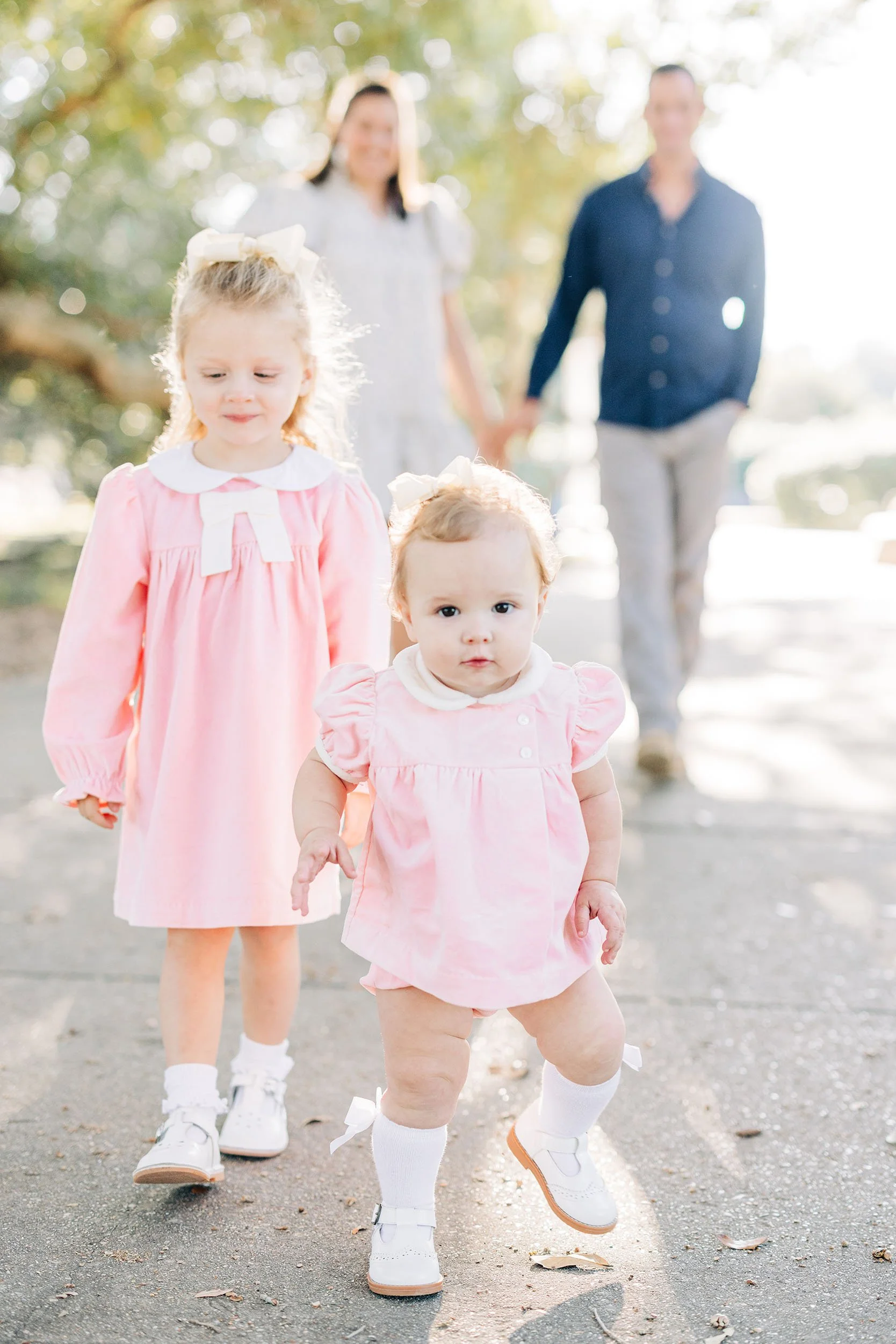 A group of children and two adults walking outdoors on a sunny day. The children are in the foreground, with a young girl in a pink dress and a younger girl in a matching pink outfit. The adults are in the background, holding hands and walking behind
