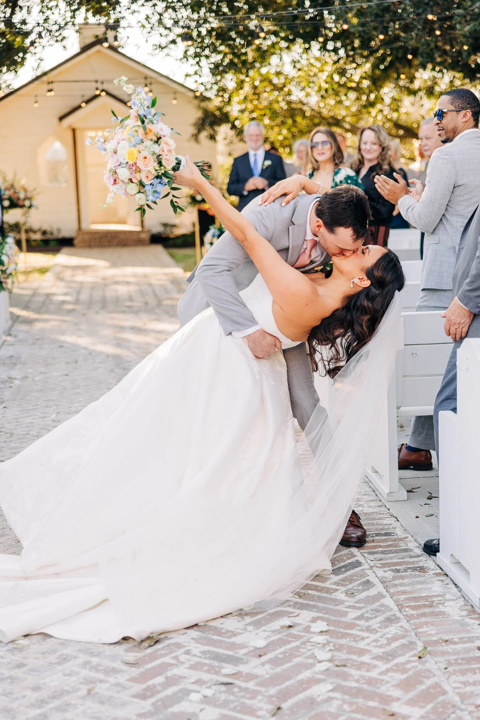 A bride and groom kissing during a wedding ceremony outdoors, with the groom bending down and holding the bride in a dip, surrounded by friends and family, with a wooden house and trees in the background.