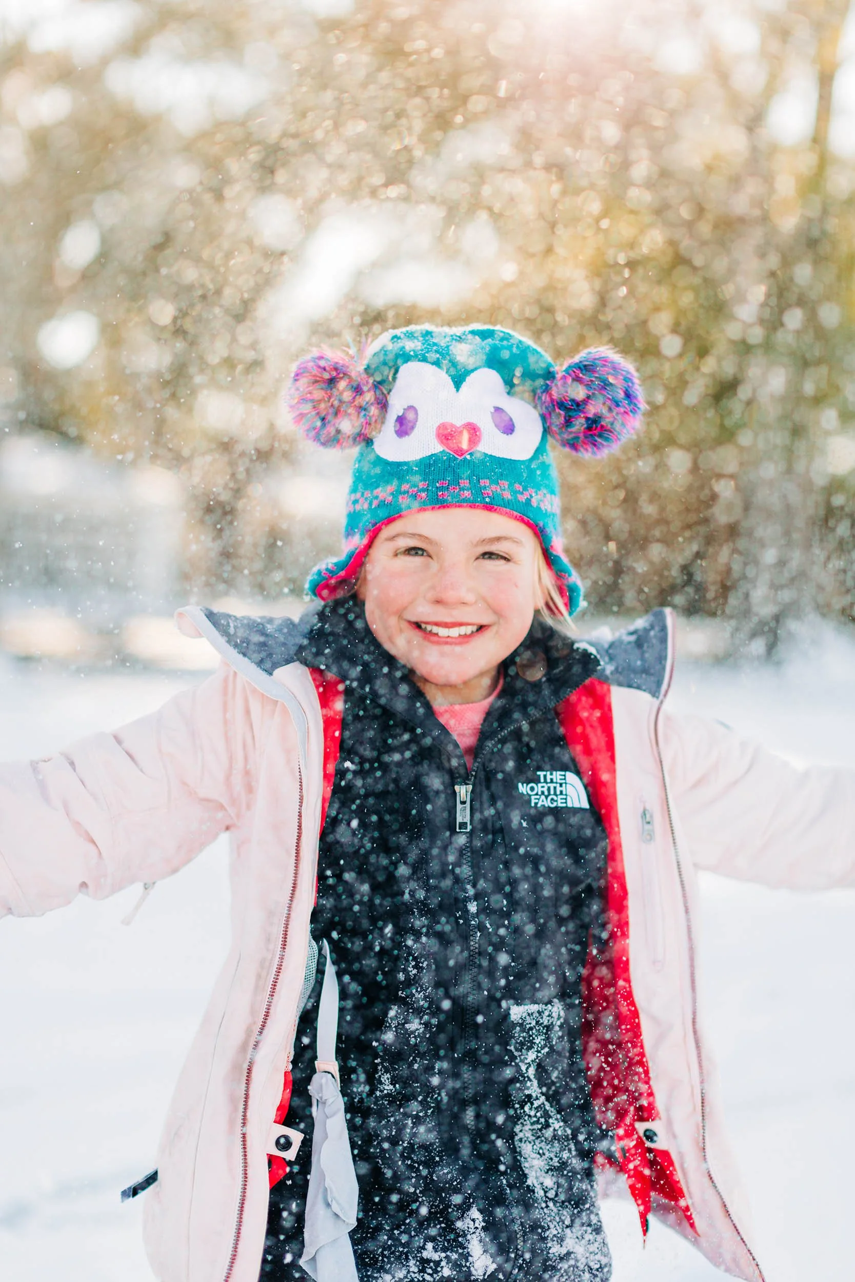 Young girl smiling outdoors in a snowy winter setting, wearing a colorful knit hat with pom-poms and a pink winter coat.