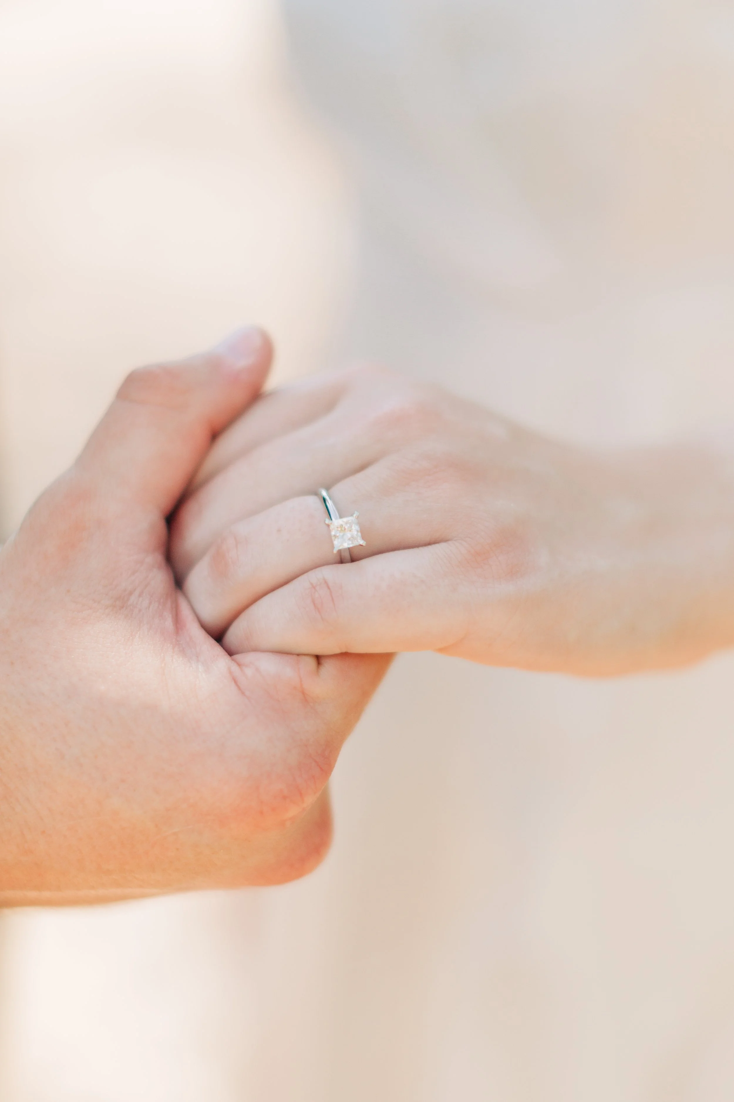 A close-up of one hand touching another, wearing a diamond engagement ring on finger, with a blurred background.