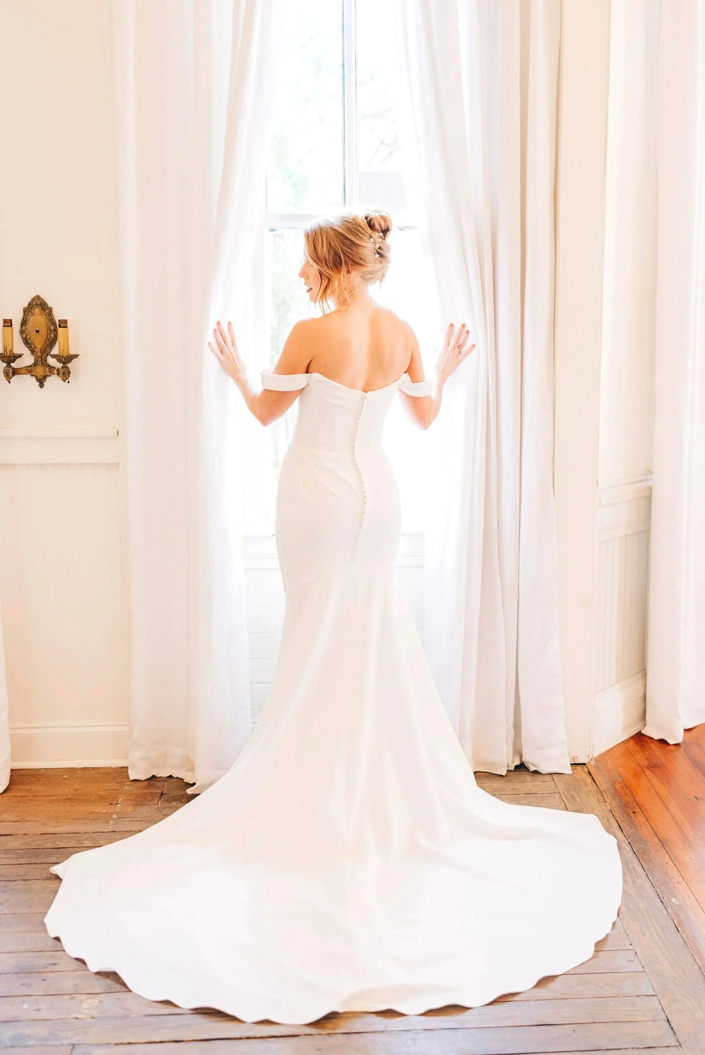 baton rouge bridal portraits, bride standing in window touching curtains with both hands, train of dress showing,  bridal gown sleeves drop over shoulders