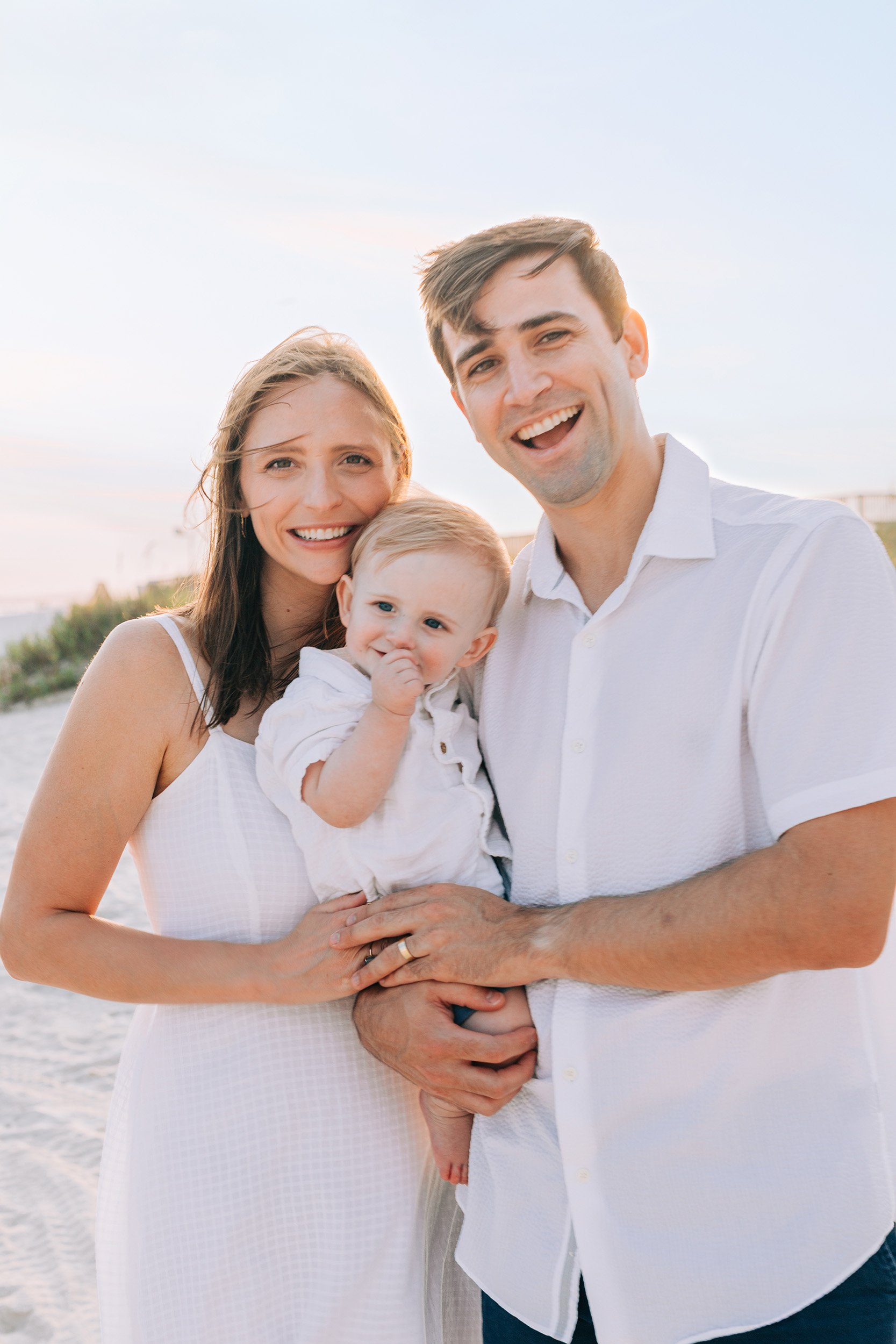 A happy family of three, a woman, a man, and a baby, standing together on a beach at sunset, smiling at the camera.