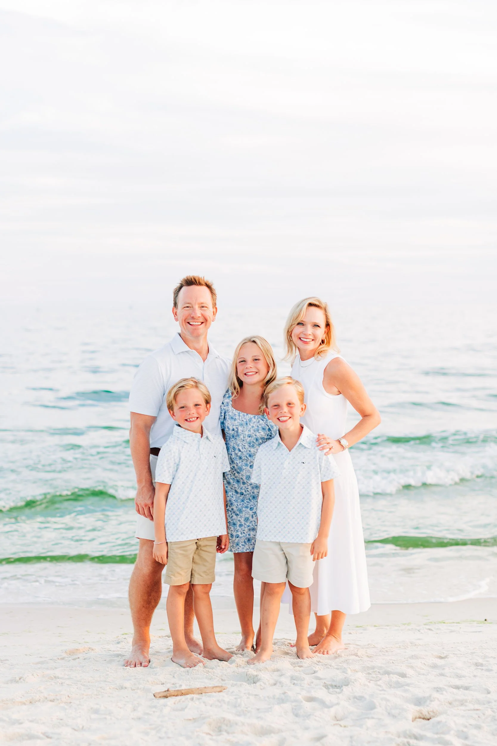 A family of seven smiling at the camera on a beach, with ocean waves and a cloudy sky in the background.