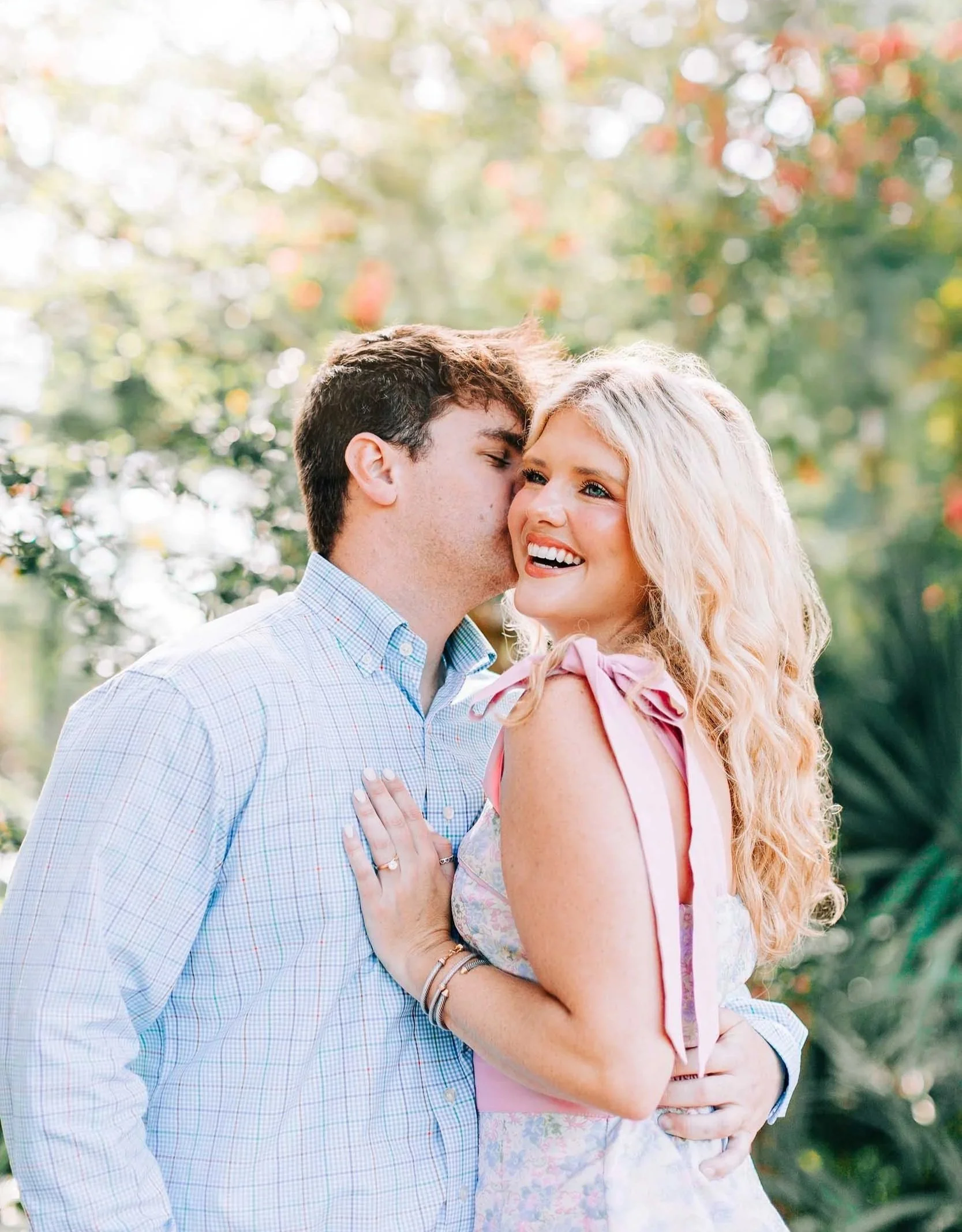 A young man and woman are outdoors, smiling and sharing a tender moment; the man is whispering into the woman's ear, and she is smiling happily.