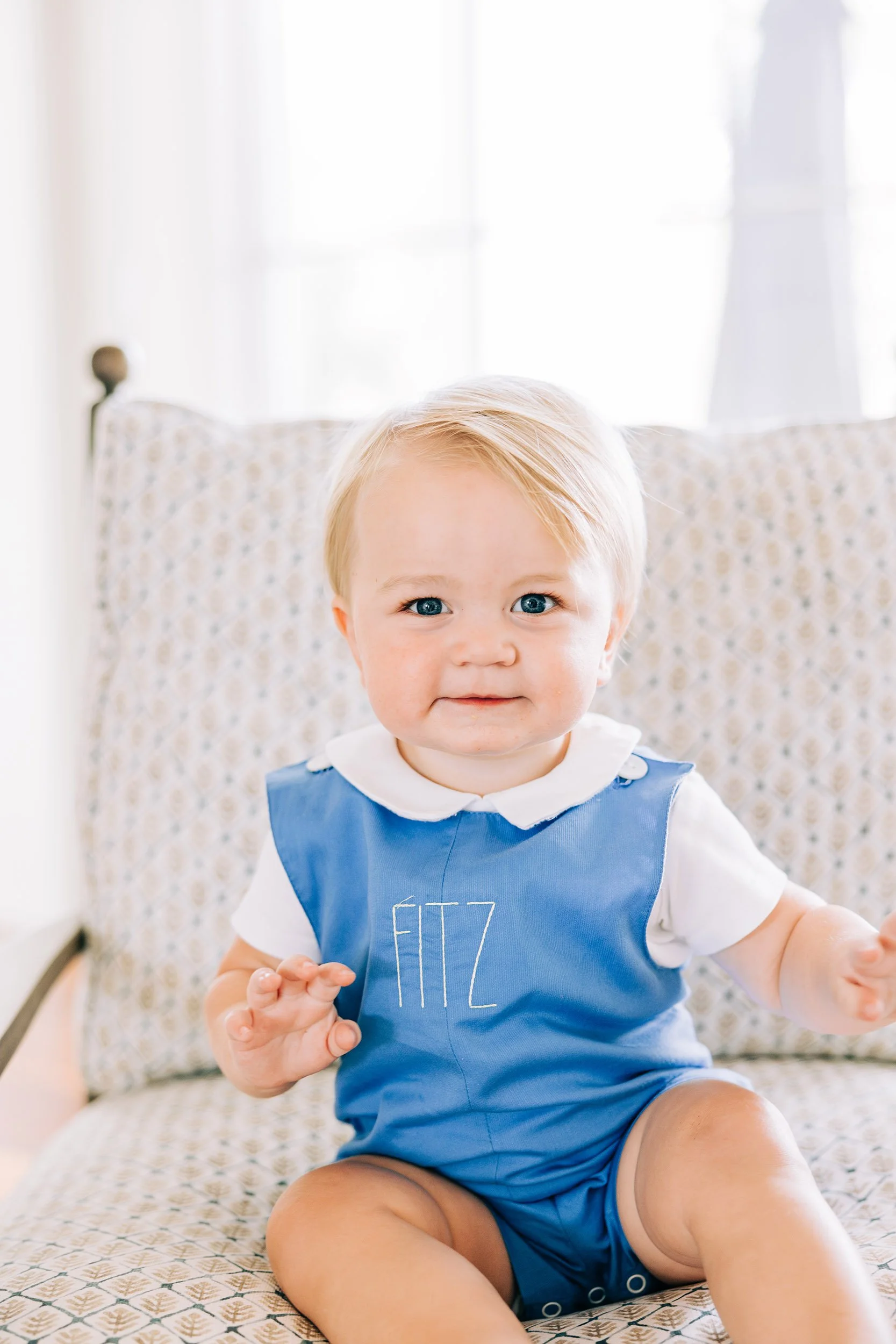 A young boy with blonde hair and blue eyes sitting on a patterned armchair, wearing a blue smock with the word "FIZ" embroidered on it, and a white shirt underneath, smiling at the camera in a bright room with white curtains in the background.