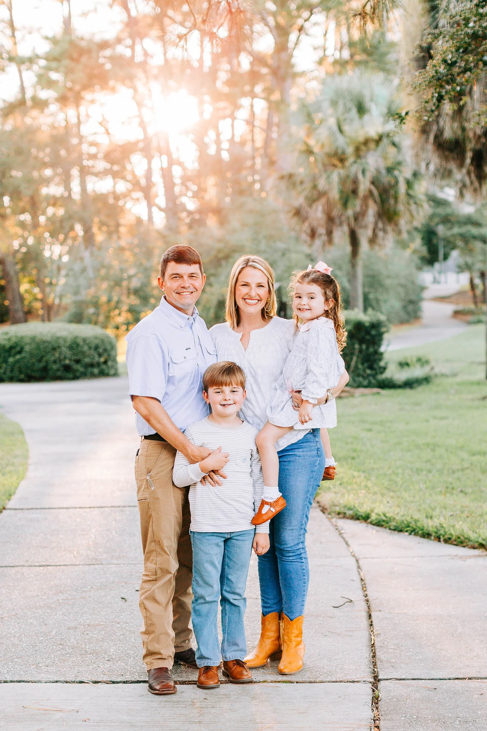 A family of five posing outdoors on a sidewalk with trees in the background during sunset. The dad and mom stand together, with their children in front and one daughter being held by the mom. They are smiling and dressed casually.