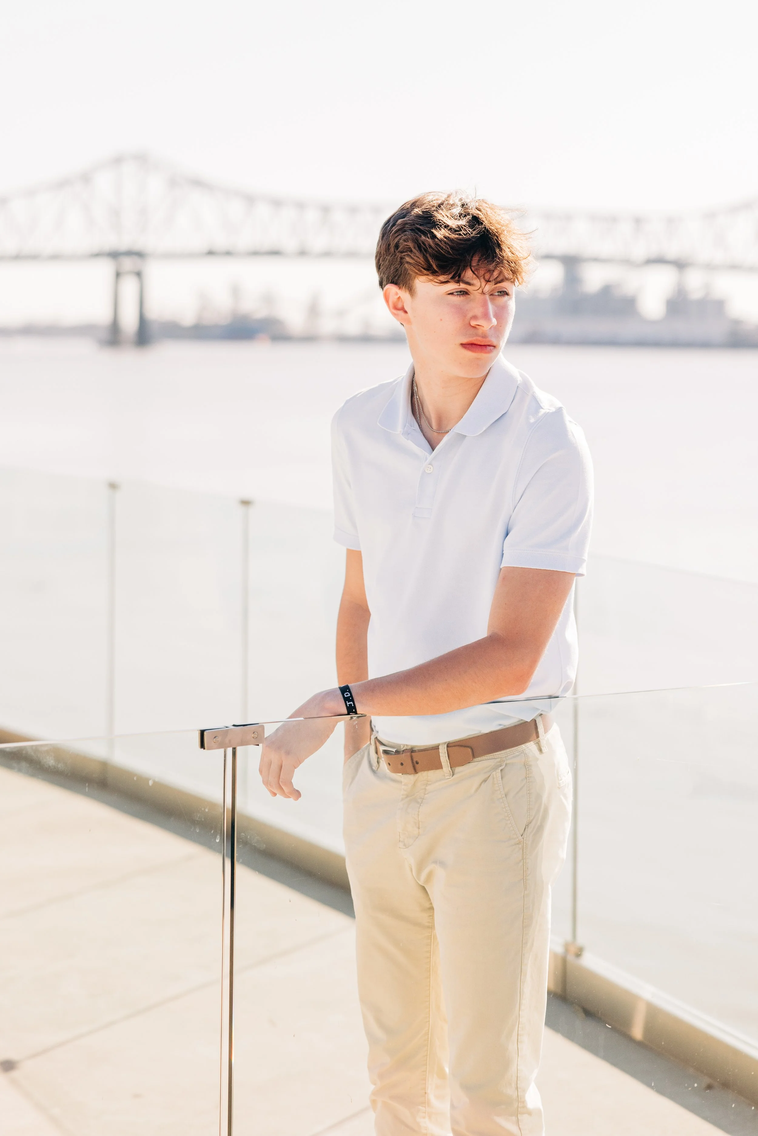 Young man with curly brown hair wearing a white polo shirt and beige pants, standing outdoors near a railing, with a bridge and city skyline in the background on a sunny day.