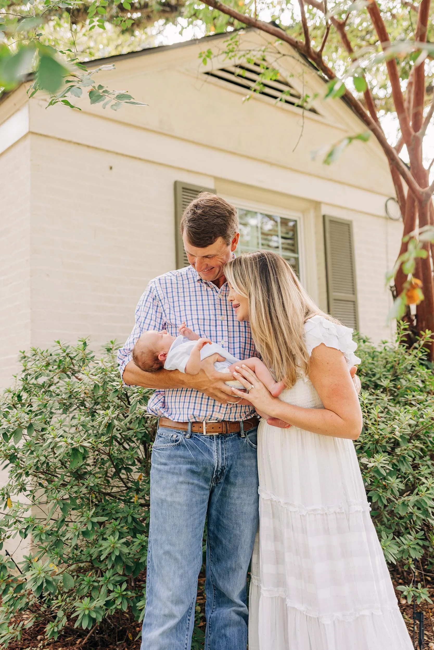 A couple holding a newborn baby outdoors in front of a house, surrounded by greenery and trees, during daytime.