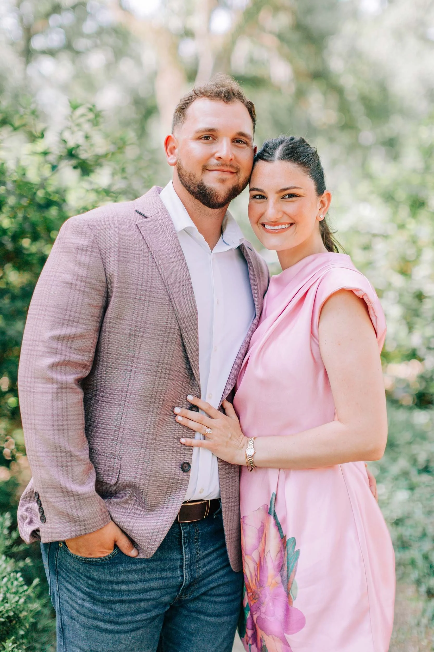 A smiling couple standing close, outdoors, in a green park, with the woman's hand on the man's chest, wearing a pink dress and the man wearing a light purple checked blazer.