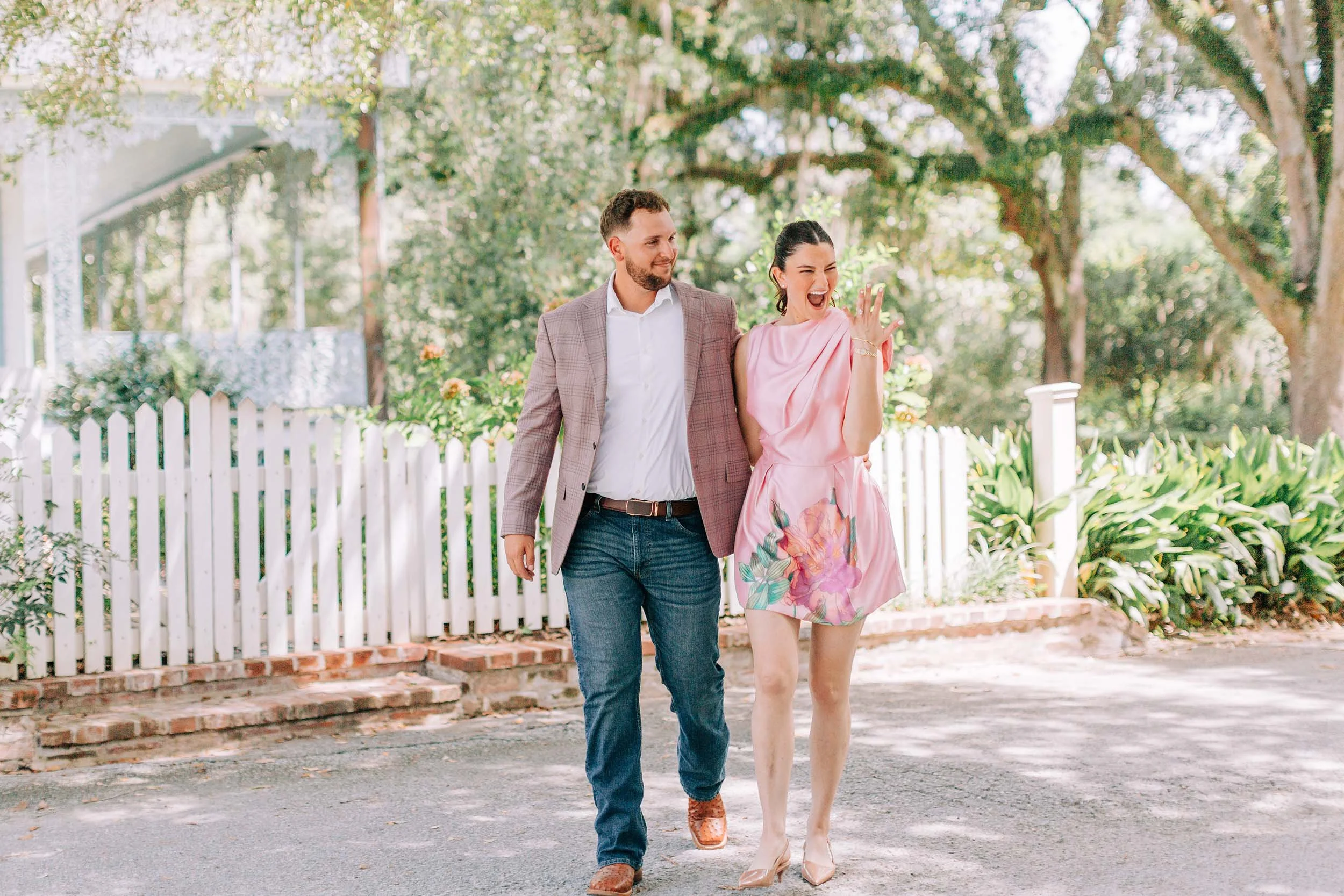 A man and woman walking together outdoors through a garden area with trees, bushes, and a white picket fence, smiling and enjoying each other's company.