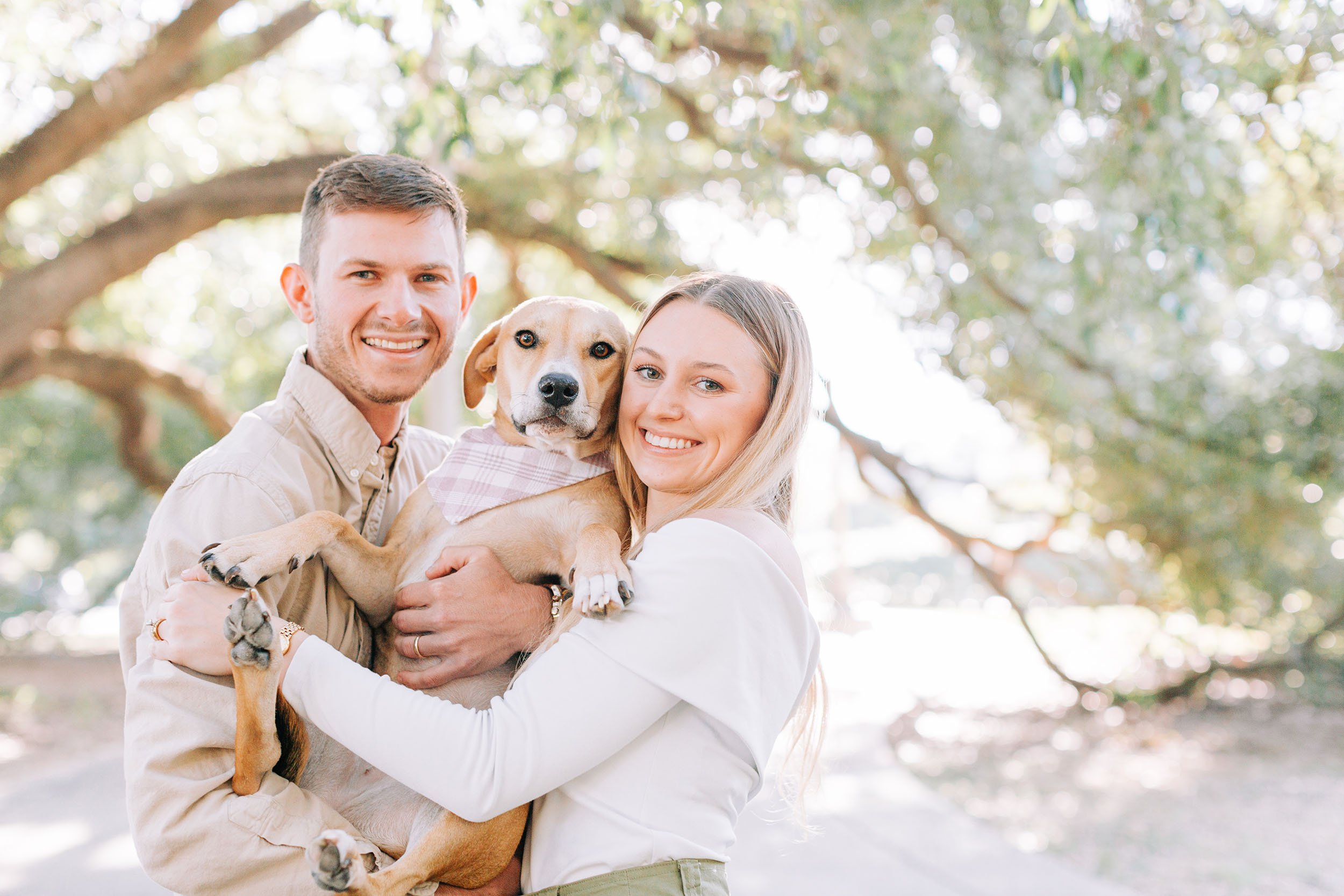 A young couple holding a dog outdoors in a park with sunlight filtering through the trees.