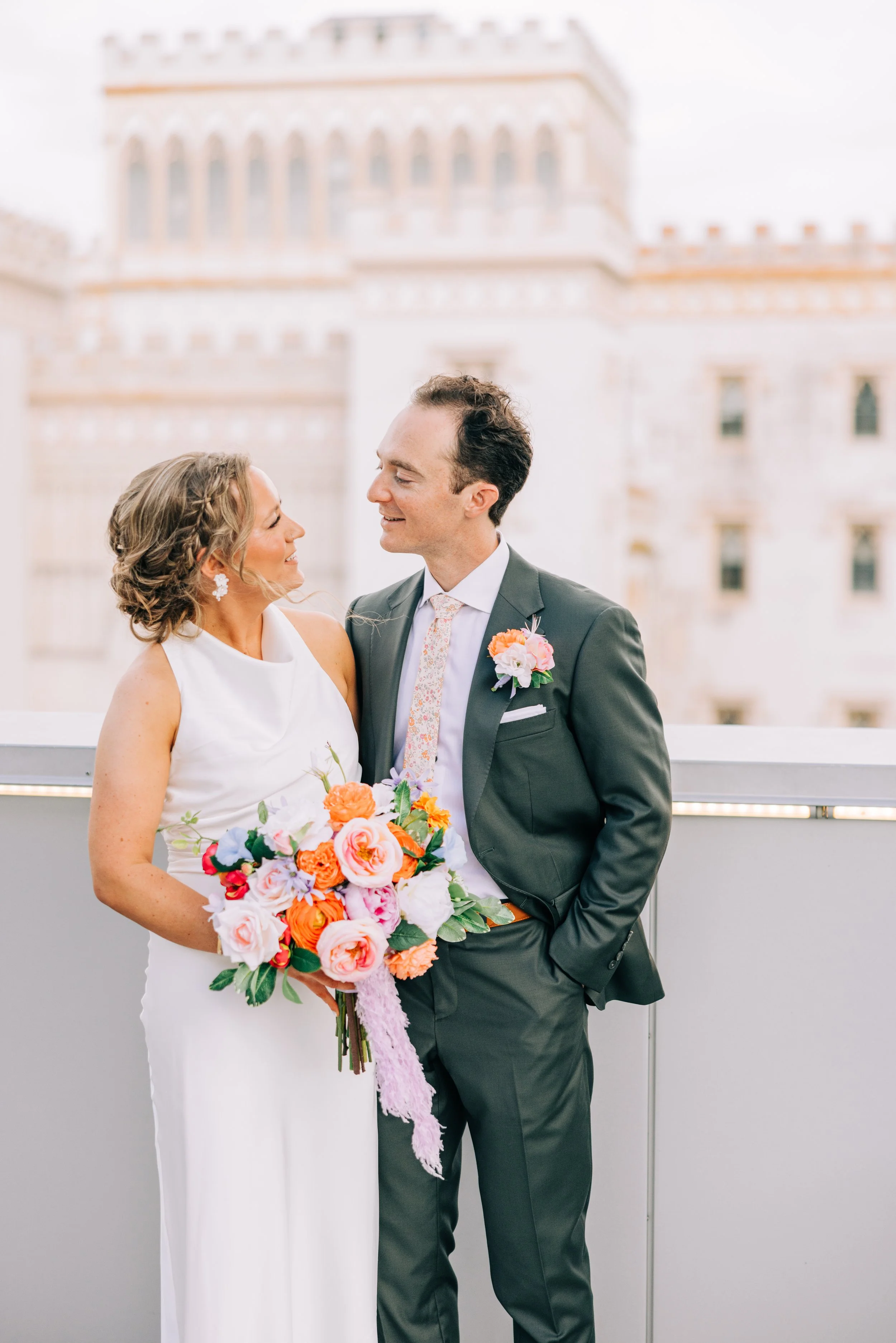 A bride and groom smiling at each other during their wedding, with a large historic building in the background