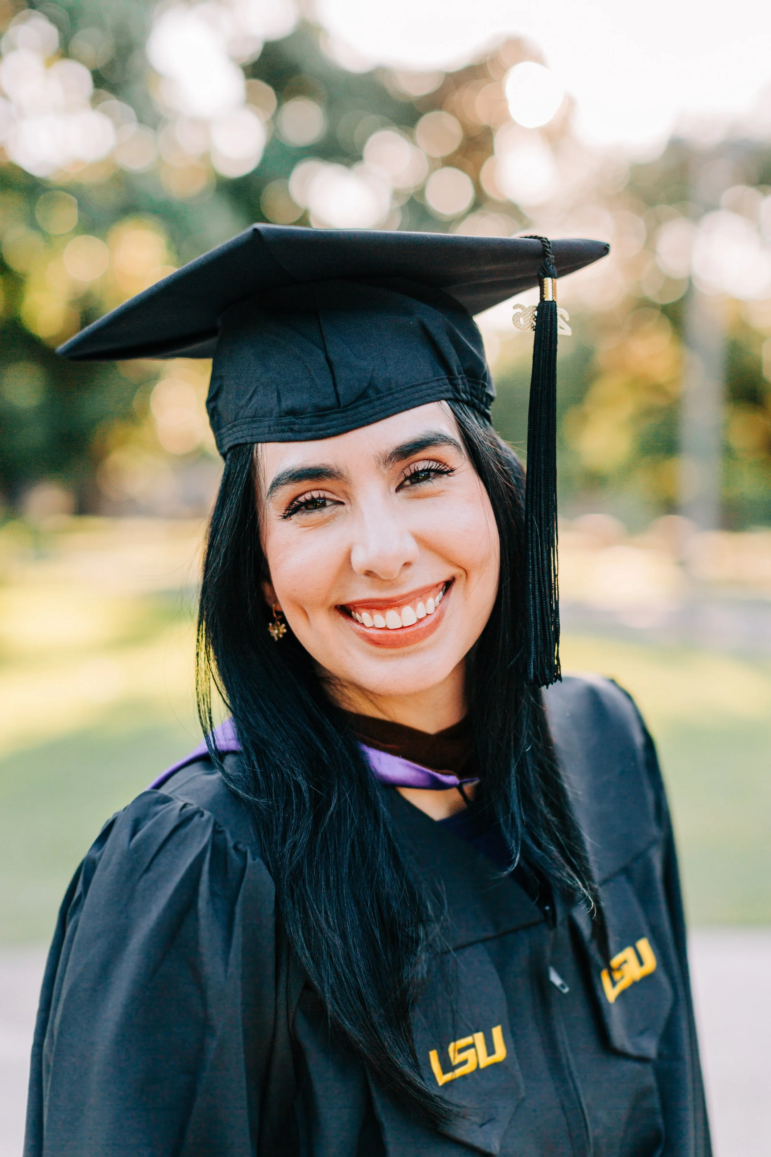 A smiling woman with dark hair in graduation regalia, wearing a black mortarboard cap with tassel, outdoors with blurred trees and bokeh sunlight in the background.