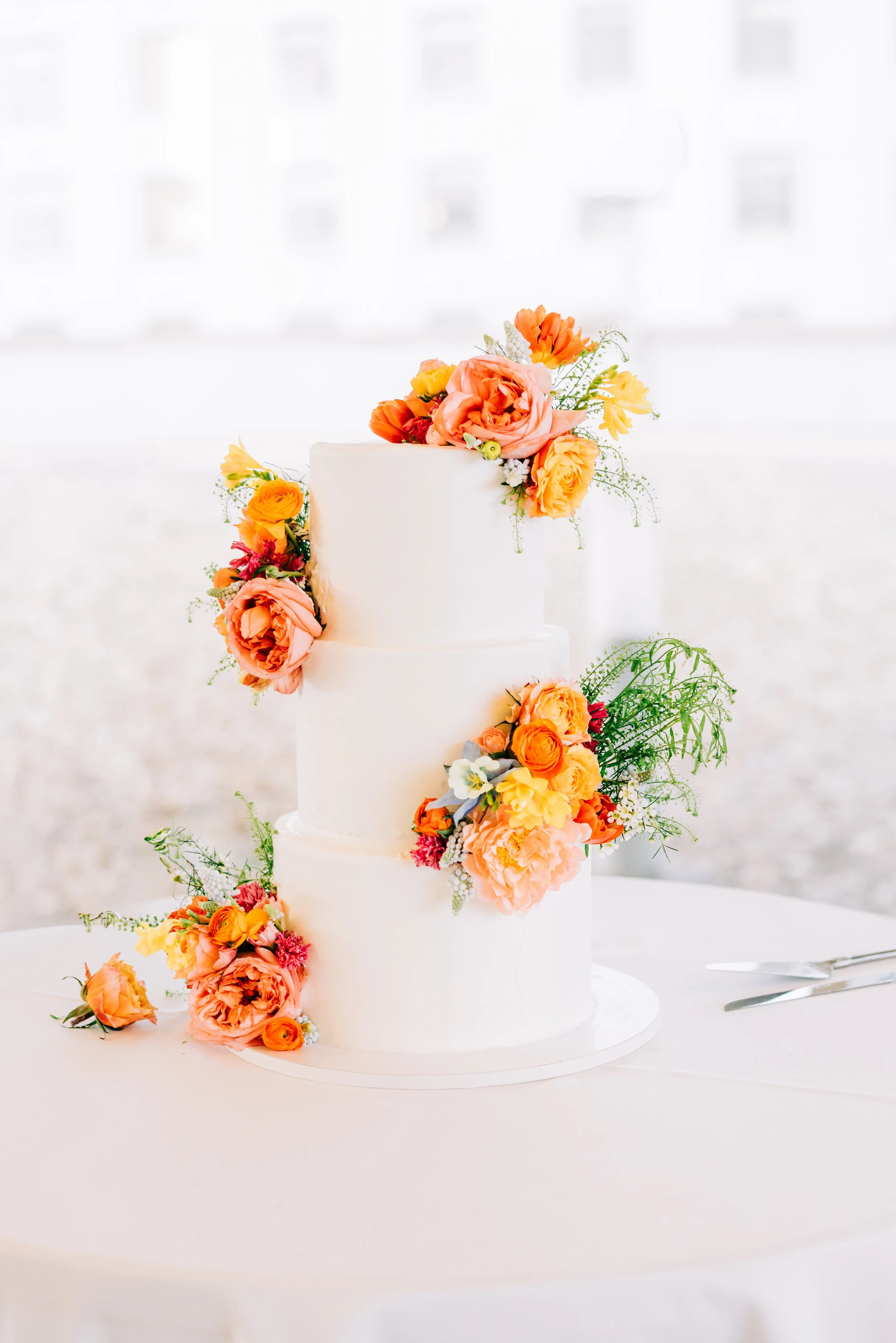 A white wedding cake decorated with cascading flowers in shades of peach, orange, and yellow, with some greenery, on a white tablecloth.