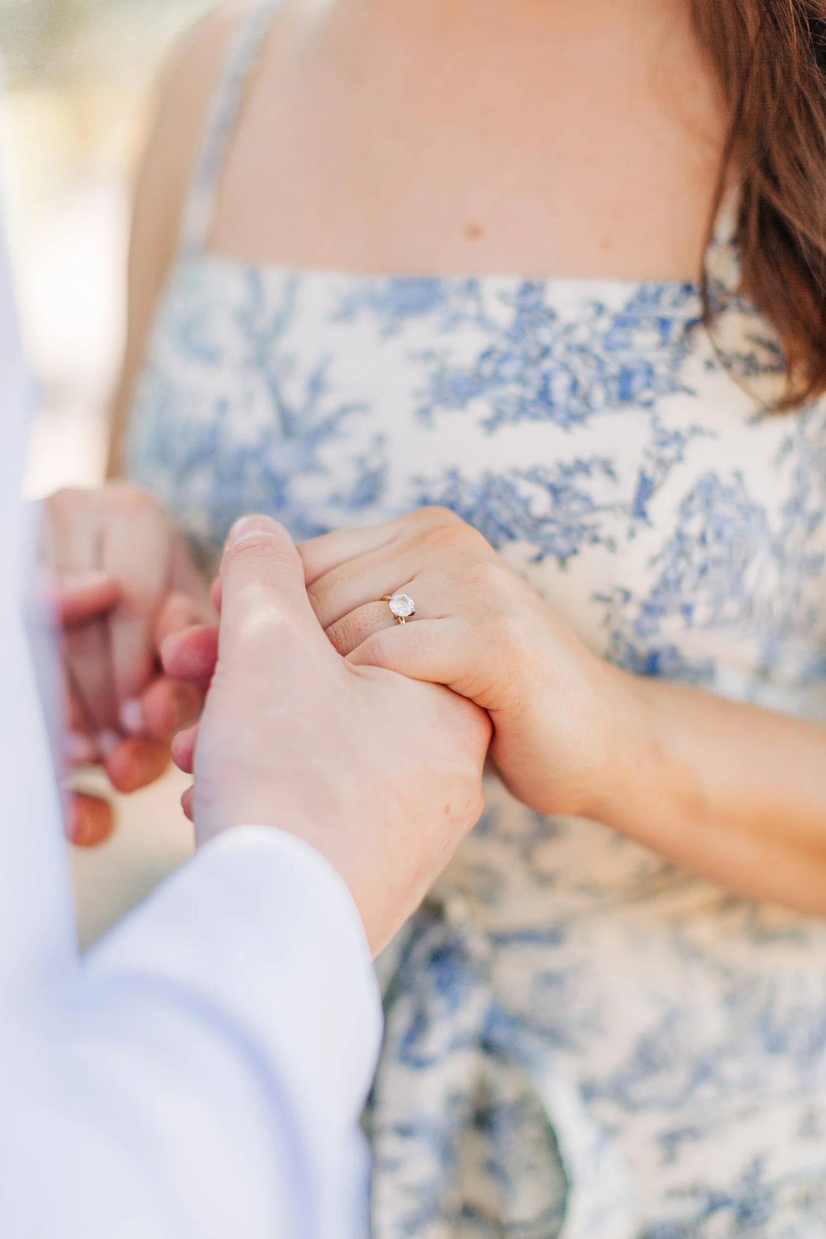 Close-up of hands holding each other; a woman wearing a wedding ring on her left hand. She is wearing a blue and white patterned dress. The background is blurred.